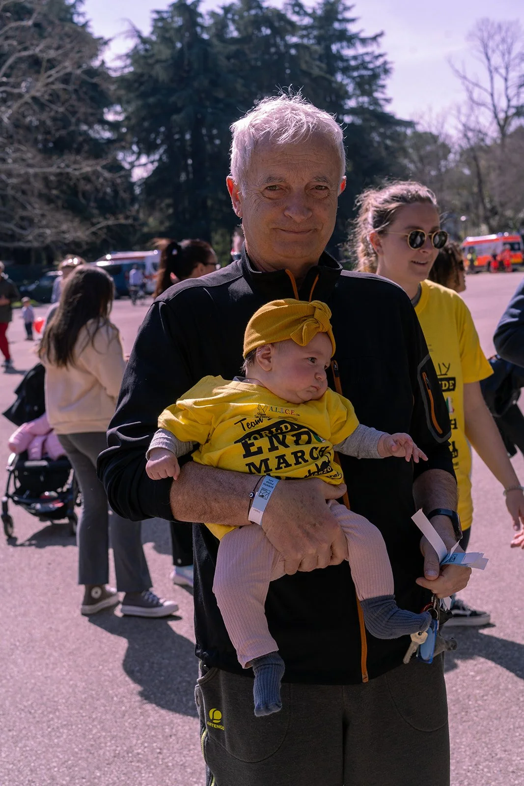Uomo che tiene in braccio un bambino, entrambi indossano magliette gialle con scritte, in un evento all'aperto con molte persone sullo sfondo e alberi.