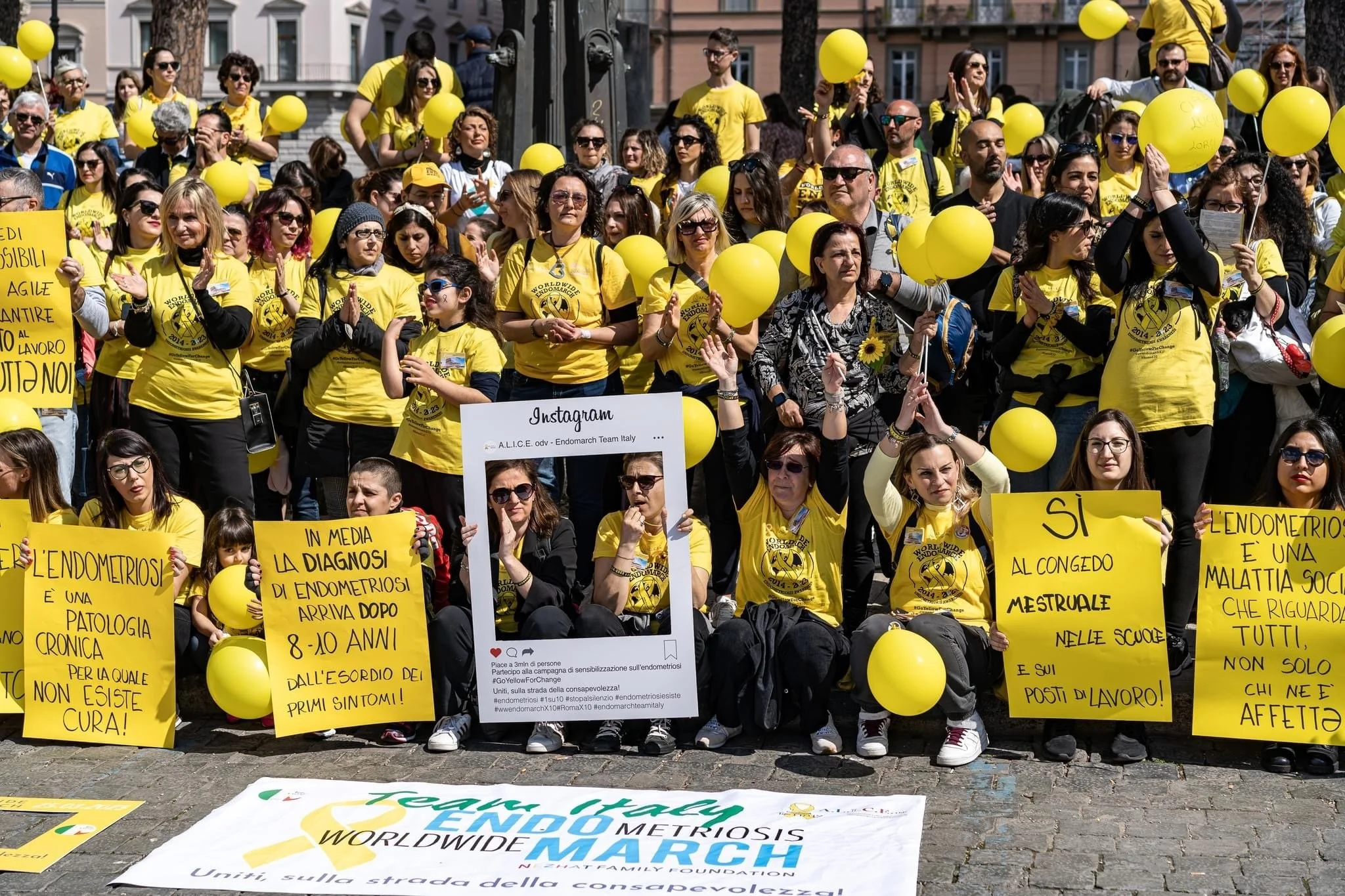 Un gruppo di persone che partecipano a una manifestazione, indossano magliette gialle con logo di Endometriosi e tengono palloncini gialli. Alcune persone tengono cartelli gialli con messaggi informativi sulla endometriosi.