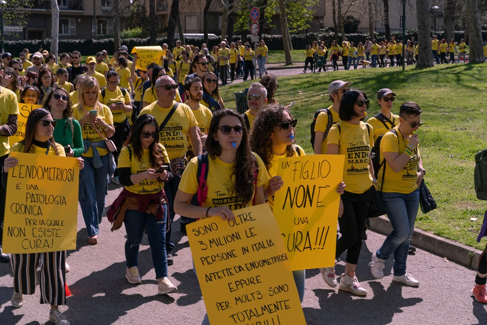 Manifestanti con magliette gialle e cartelli di protesta che camminano in una marcia, alcuni con bottiglie d'acqua in bocca, in un parco.