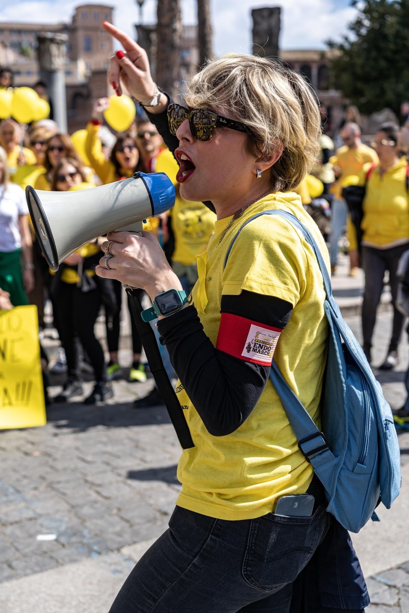 Donna con occhiali da sole e t-shirt gialla con bracciale rosso che parla attraverso un megafono durante una manifestazione in strada, circondata da altre persone con magliette gialle.
