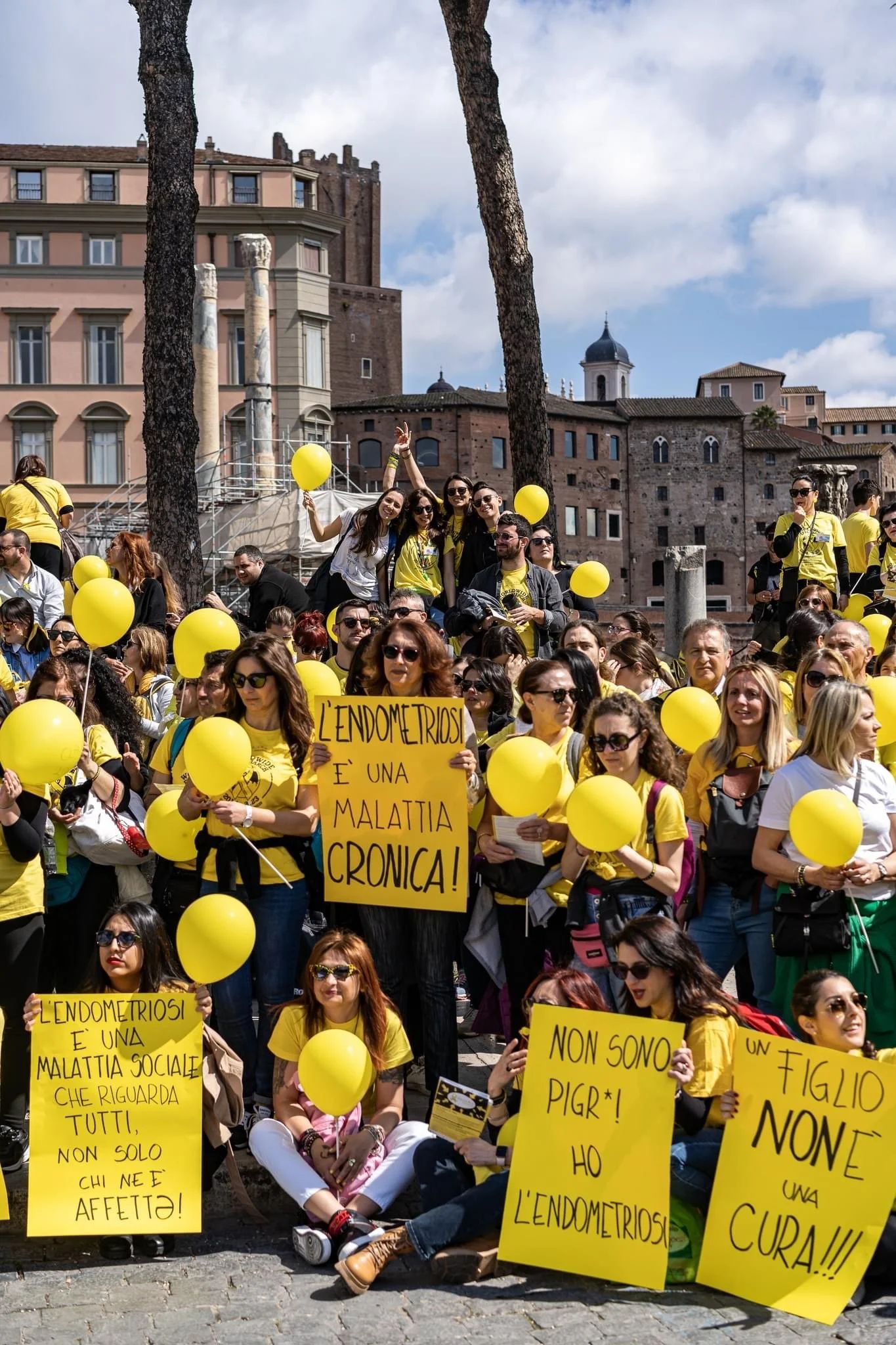 Gruppo di persone partecipano a una manifestazione, alcuni tengono palloncini gialli e cartelli con scritte in italiano, in una piazza con edifici storici sullo sfondo.