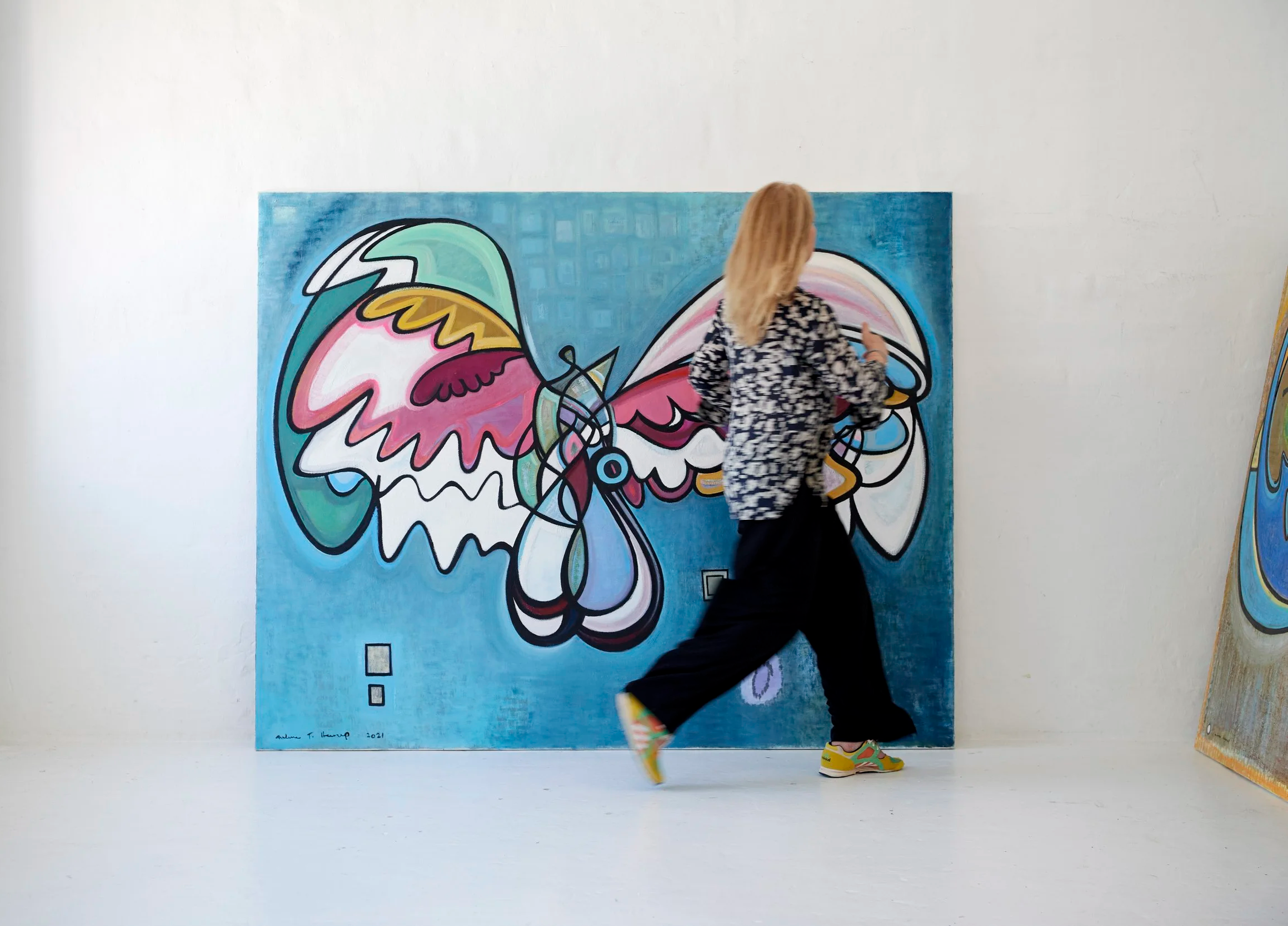 A woman with blonde hair walking in front of a colorful abstract butterfly painting in an art gallery.