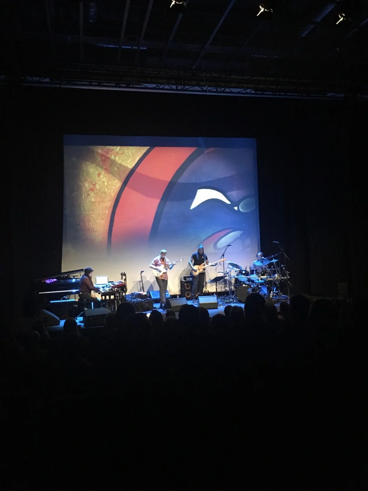 A band performing on stage in a dark theater with a large screen displaying abstract artwork behind them.