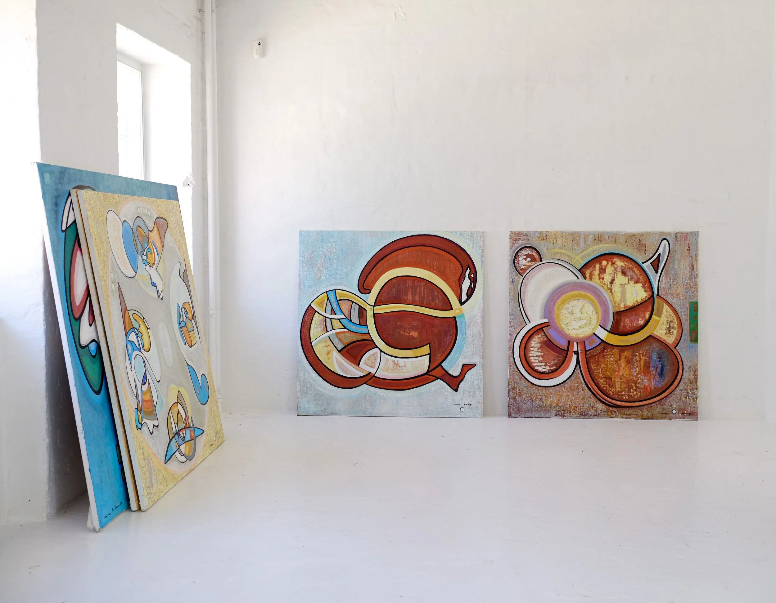Three abstract paintings with vibrant, curvy lines and geometric shapes, displayed against a white wall in an art gallery.