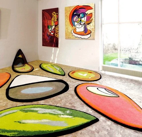 A gallery room with colorful abstract rugs shaped like peels on the floor, and two vibrant paintings on the wall. Natural light streams through a large window.
