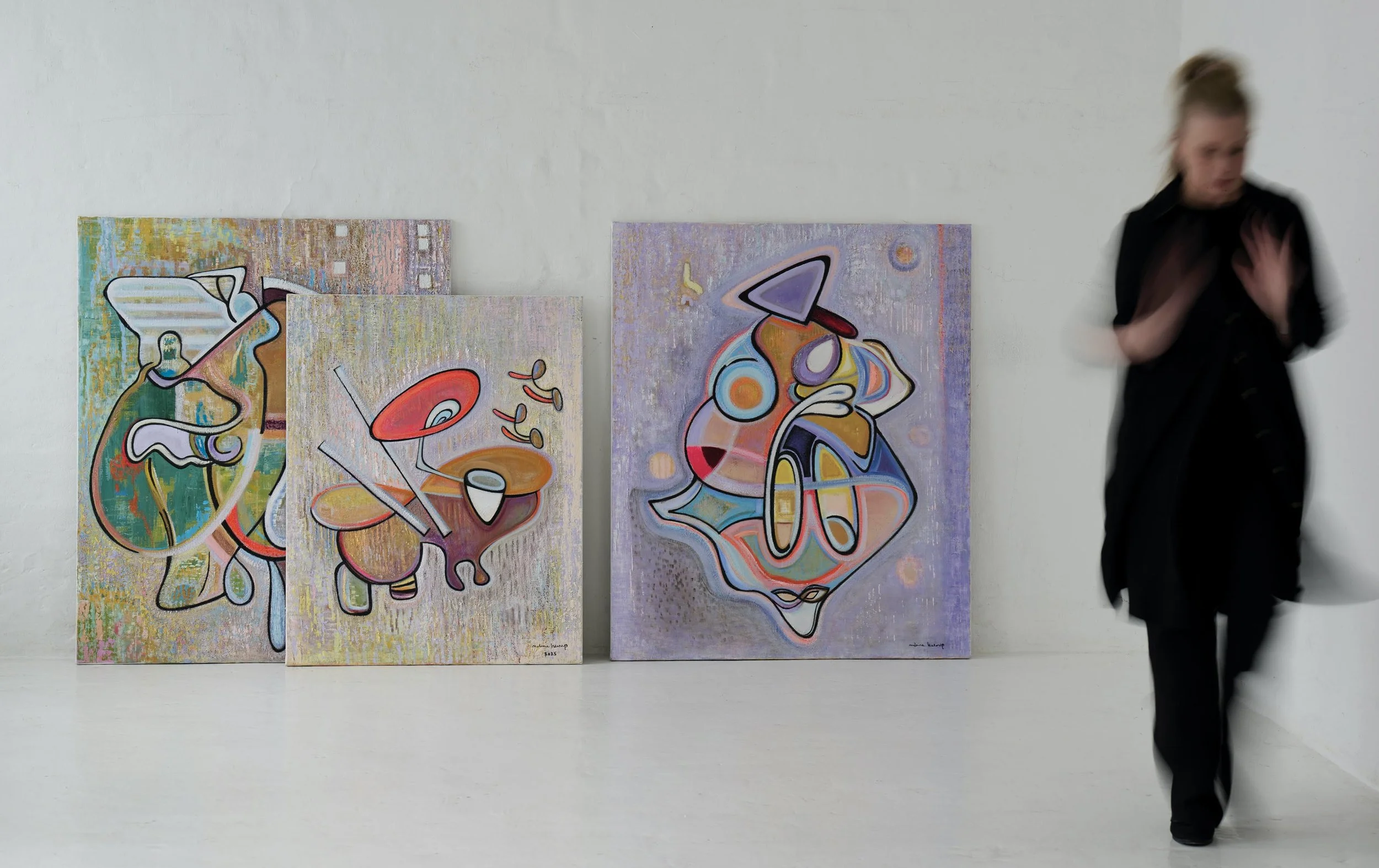 Three colorful abstract paintings leaning against a white gallery wall, with a blurred woman walking past on the right side.