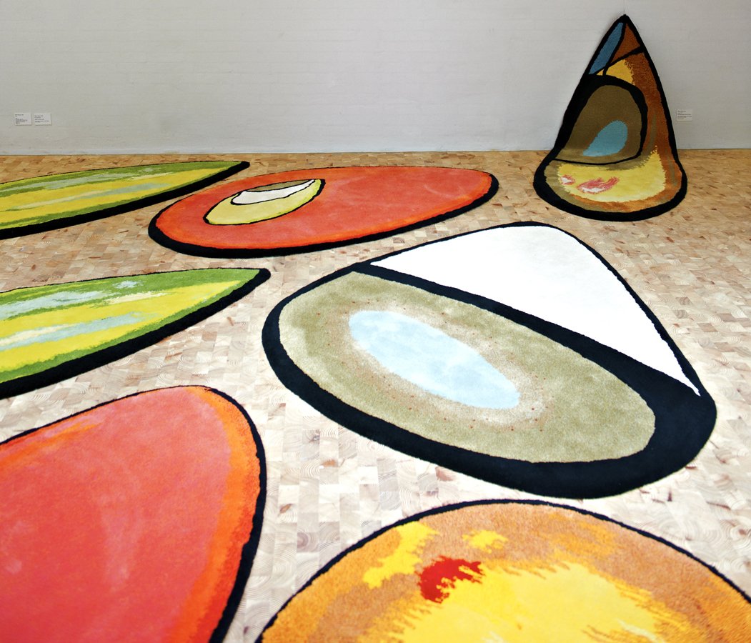 Colorful, abstract art rugs resembling fruit slices and vegetables in a gallery setting.