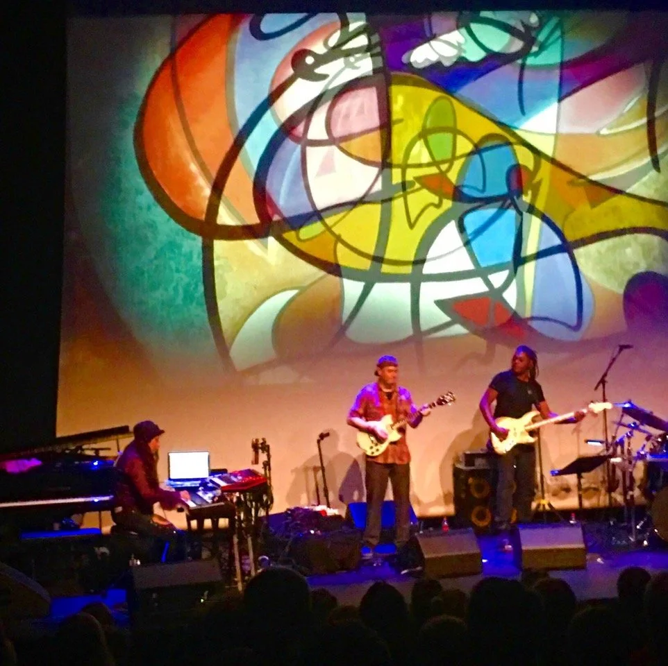A band of three musicians performs on stage with a large abstract colorful mural projected behind them.