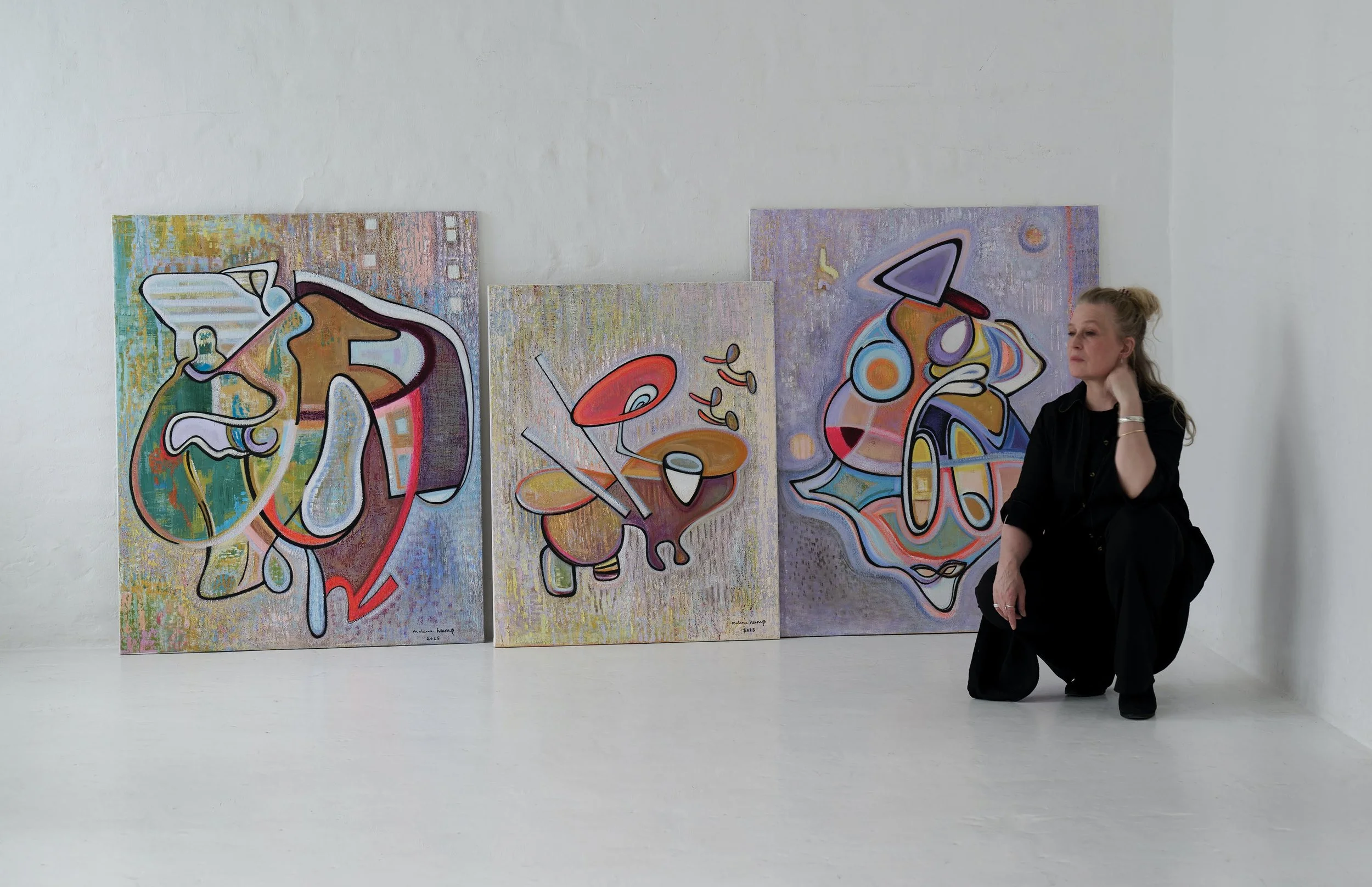 A woman with blonde hair in a bun, dressed in black, kneeling in an art gallery with three colorful abstract paintings on the wall behind her.