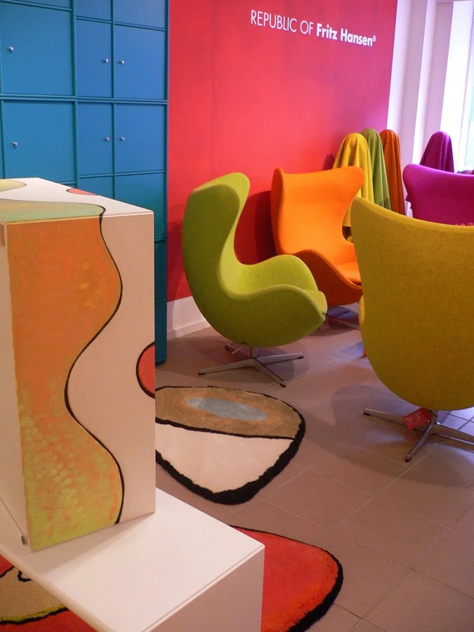 Colorful modern chairs in green, orange, yellow, and pink arranged in a room with a red wall and blue lockers, with a patterned rug on the floor, and a cabinet with a colorful abstract design.