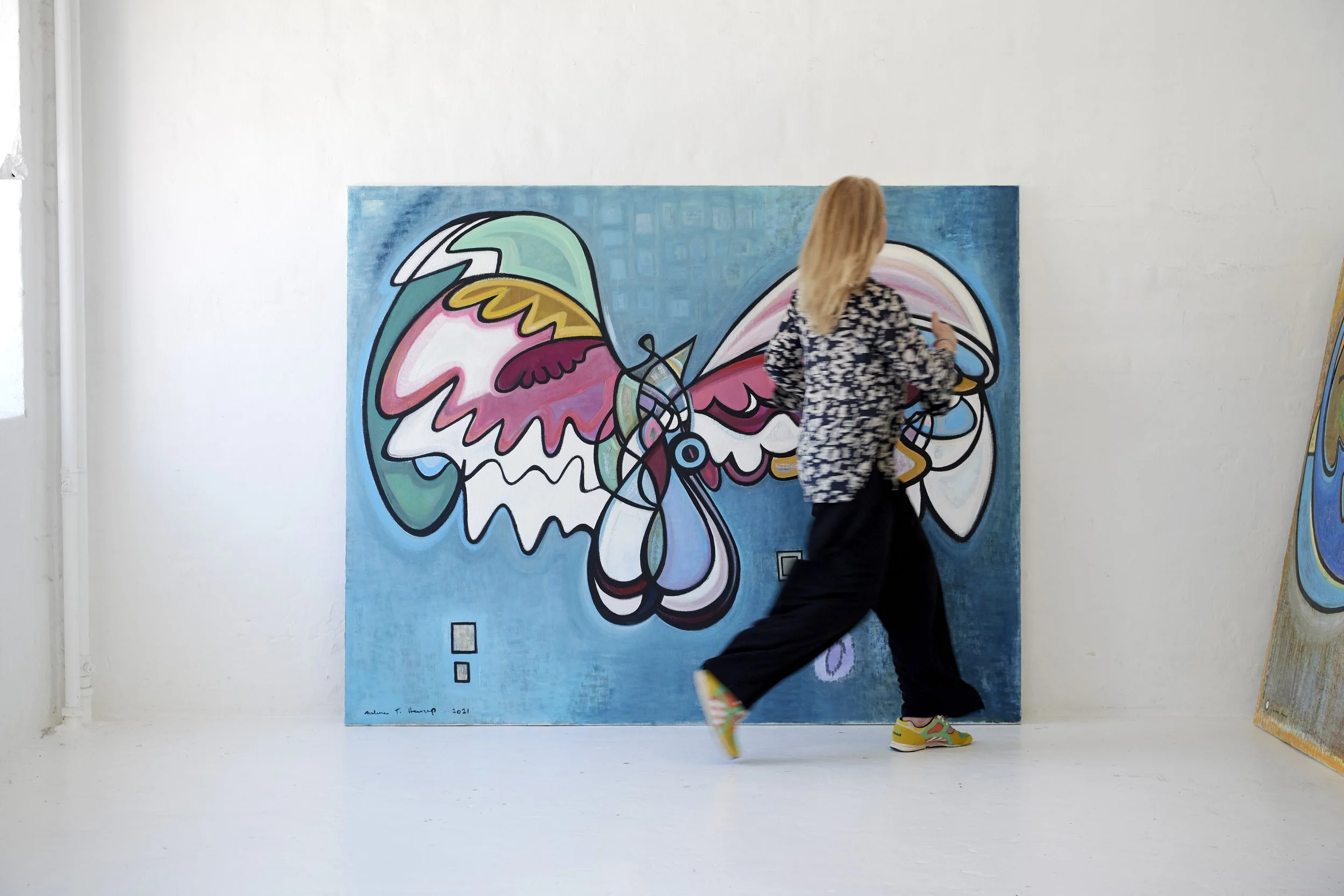 A woman with blonde hair walking in front of a colorful abstract butterfly painting in an art gallery.
