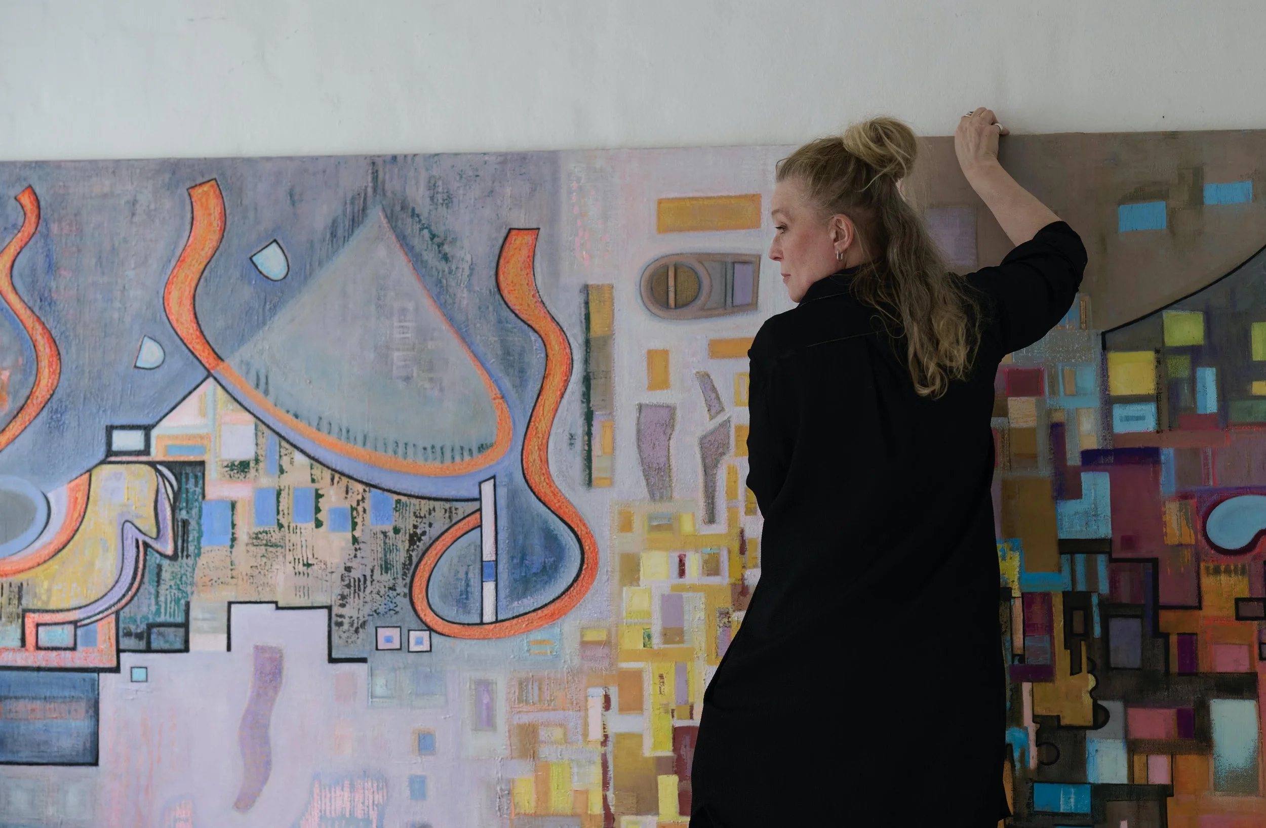 An artist with blonde hair tied up, wearing a black dress, is painting an abstract, colorful mural on a large wall.