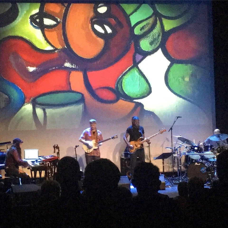 A band performing live on stage with a colorful abstract artwork projected behind them.