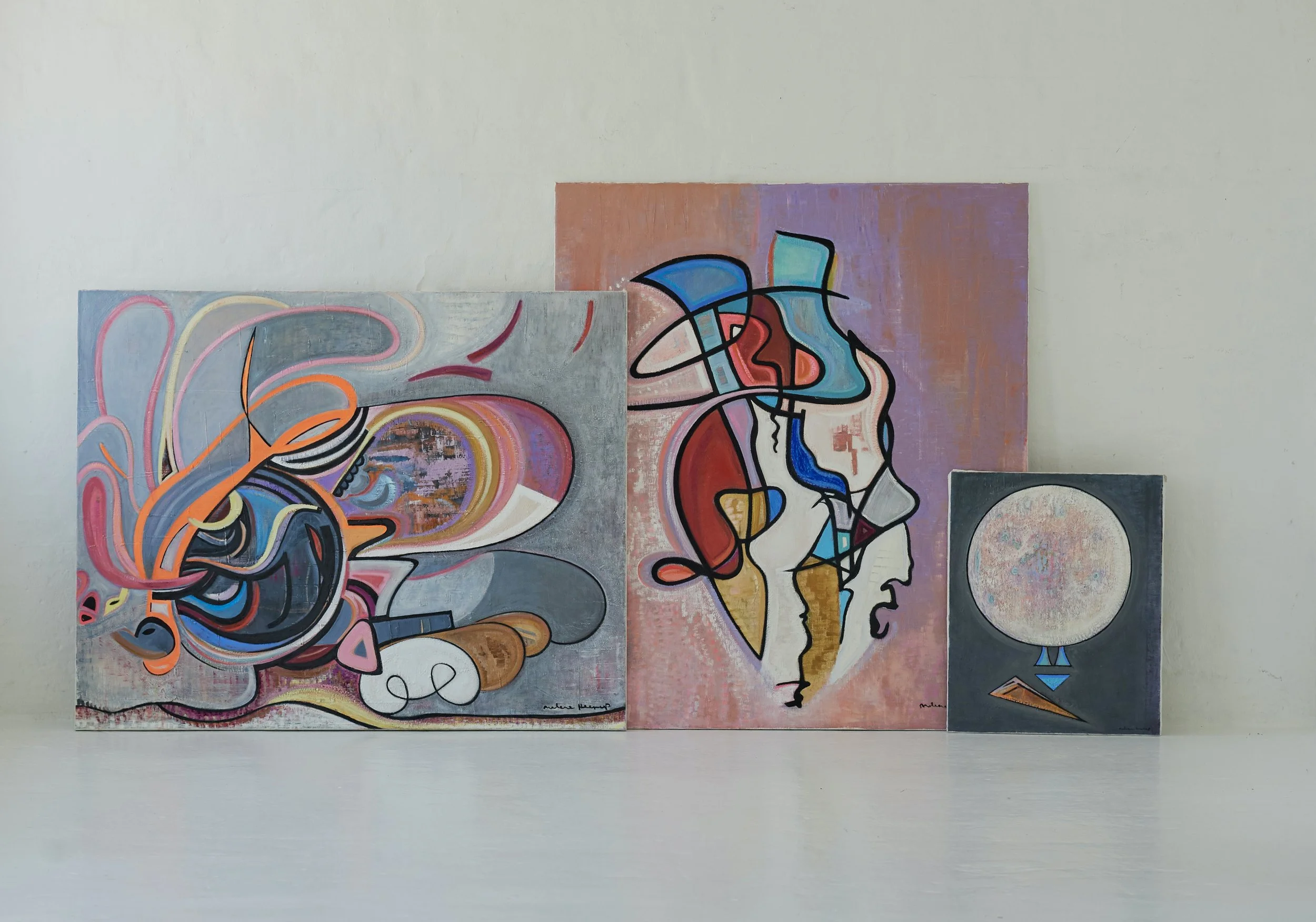 Three abstract paintings leaning against a white wall, featuring colorful shapes and lines with a mix of geometric and organic forms.