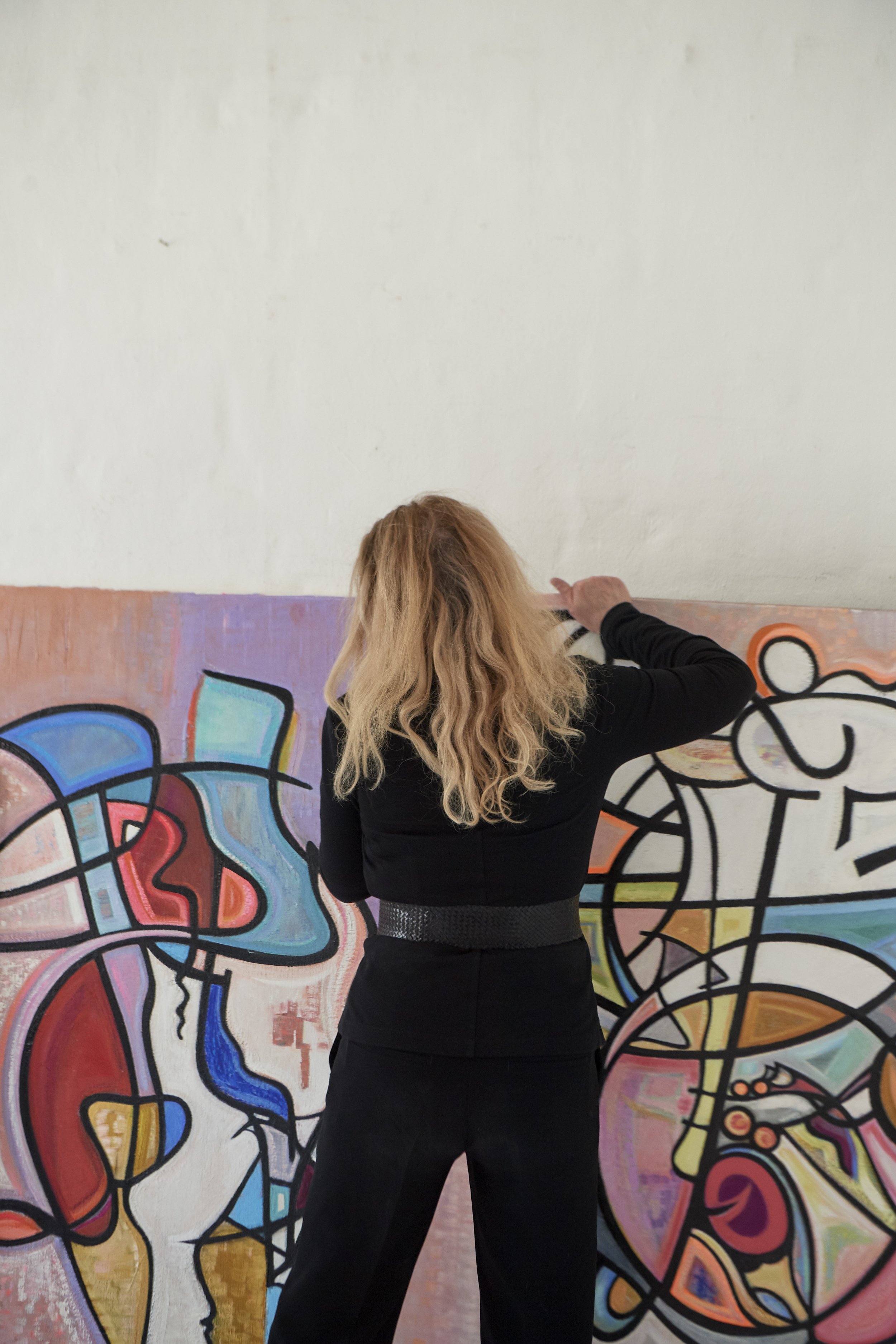 A woman with long, wavy, blonde hair wearing a black outfit, painting on a colorful abstract mural on a wall.