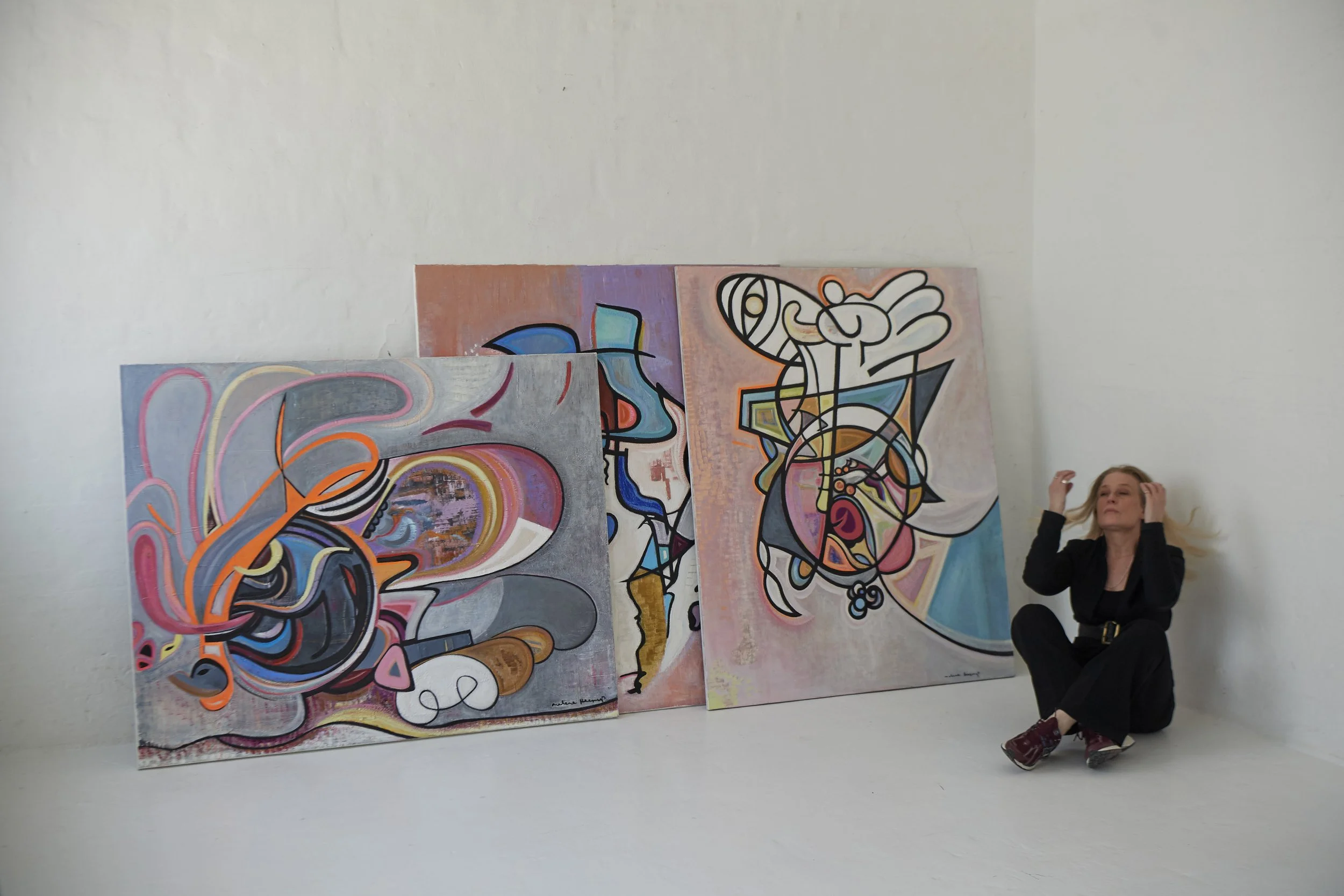 A woman sitting on the floor touching her hair, next to three large abstract paintings leaning against a white wall in the art gallery.