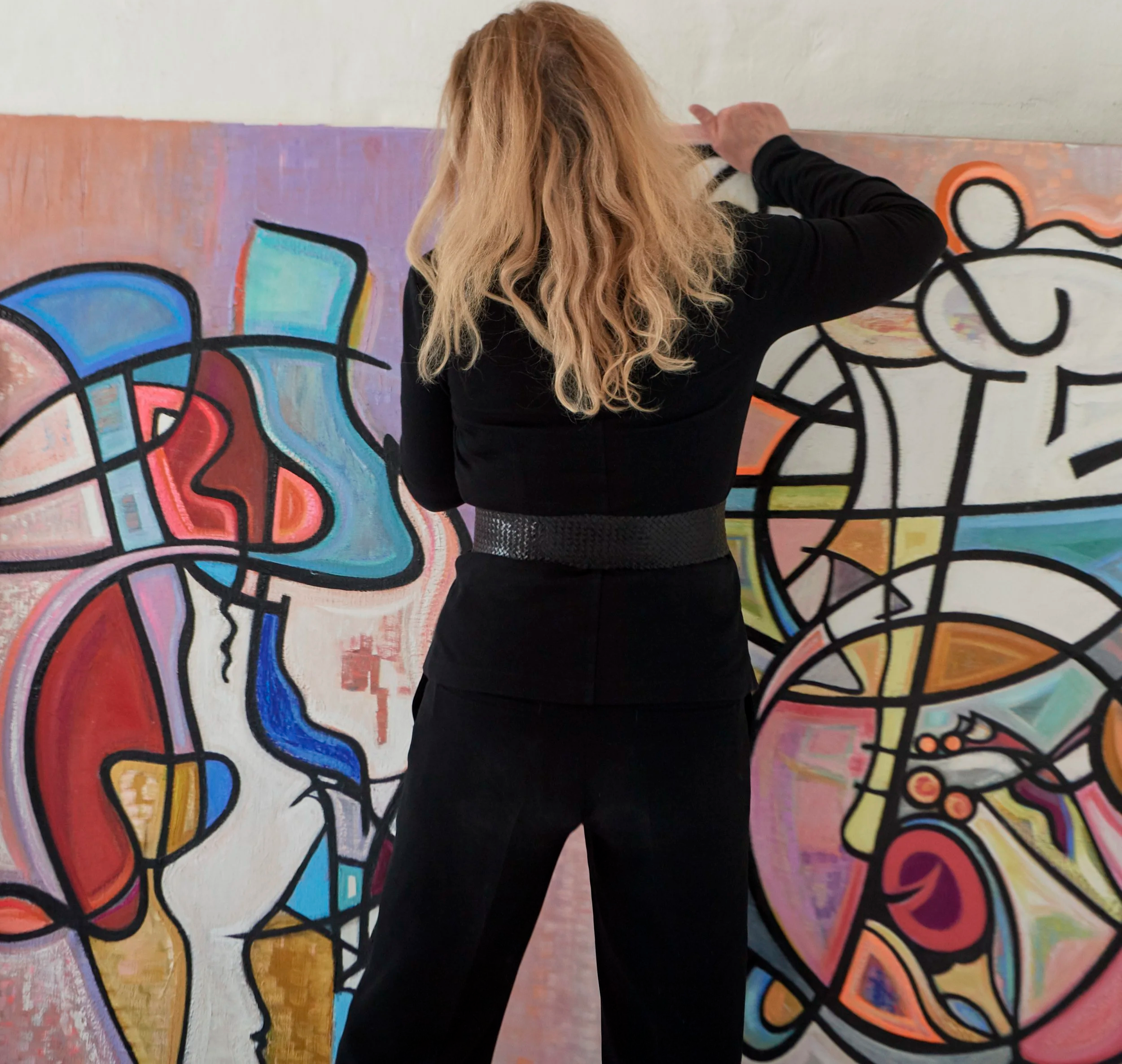 Person with long wavy blonde hair wearing black clothing standing in front of an abstract colorful mural with curved shapes and bold black outlines.