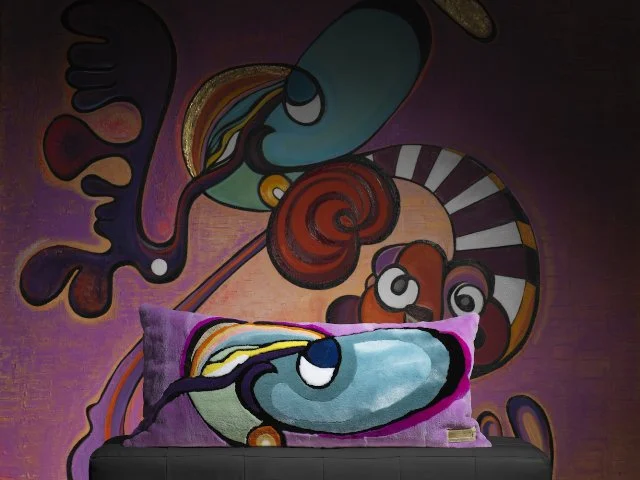 A colorful abstract mural with swirling shapes and faces, featuring bold lines and vibrant colors, behind a couch with a purple pillow that has a matching abstract design.