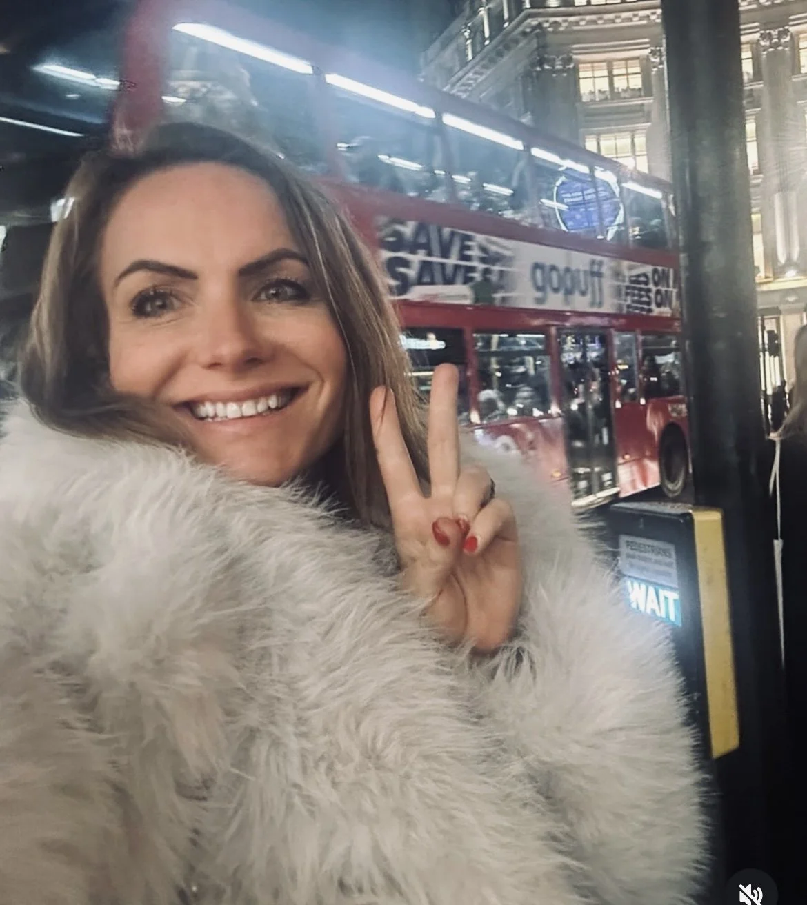 A woman smiling and showing a peace sign, standing near a bus in a city at night.