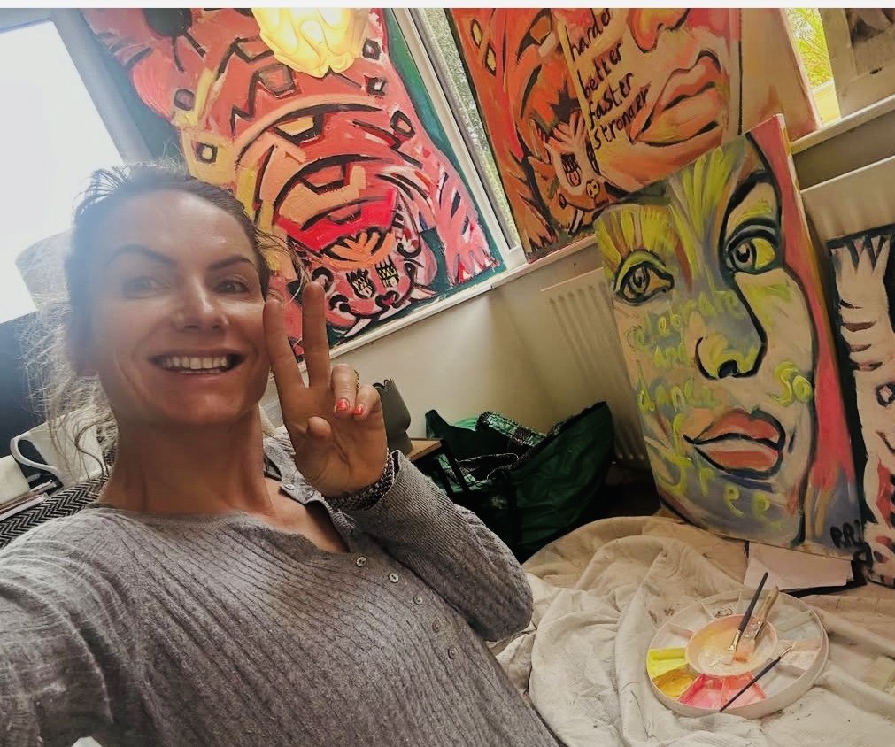 A woman smiling and making a peace sign in front of colorful, abstract paintings in an art studio.