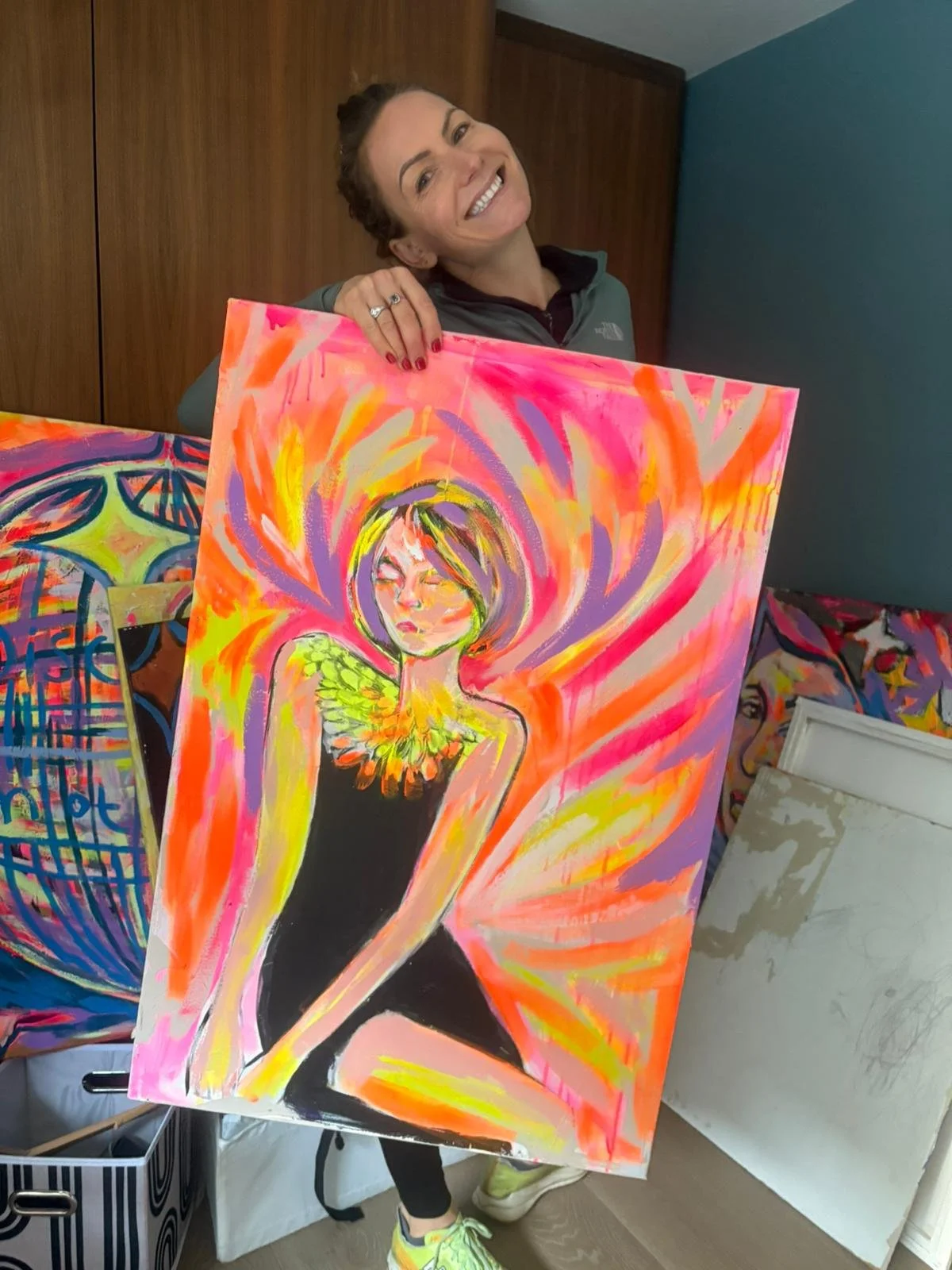 A smiling woman holding a colorful abstract painting of a woman with wings, surrounded by vibrant orange, pink, and purple brush strokes.