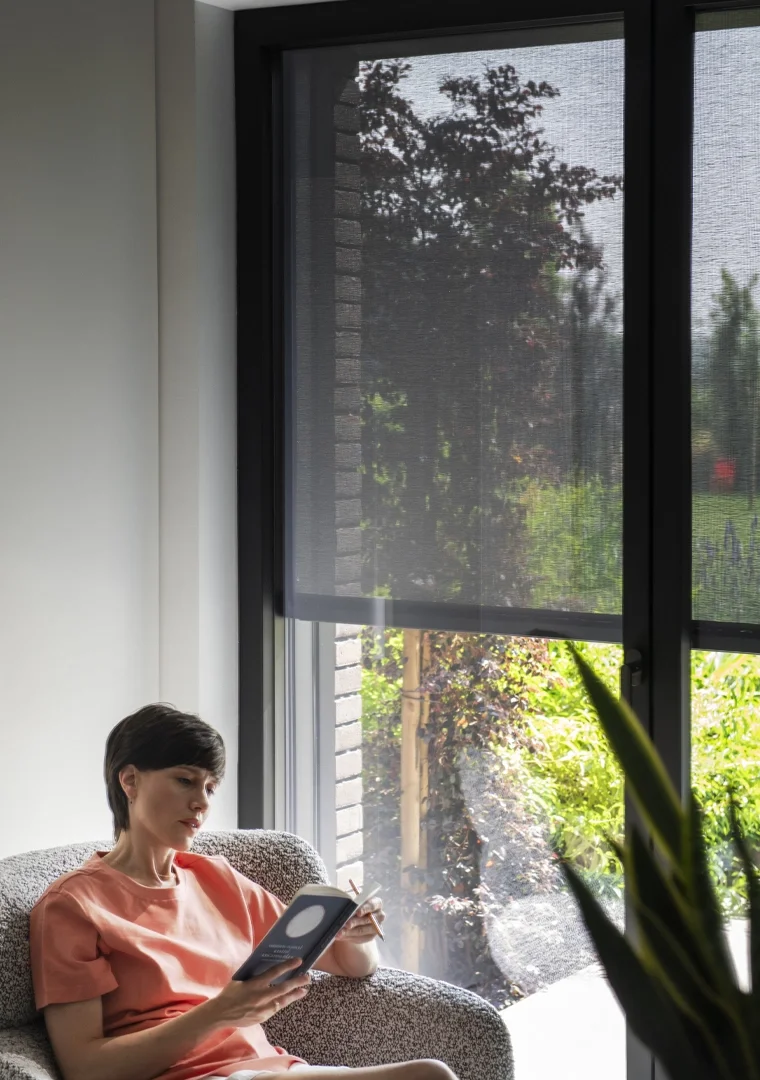 A woman sits on a gray couch near a window, reading a book with sunlight streaming in and a view of greenery outside.