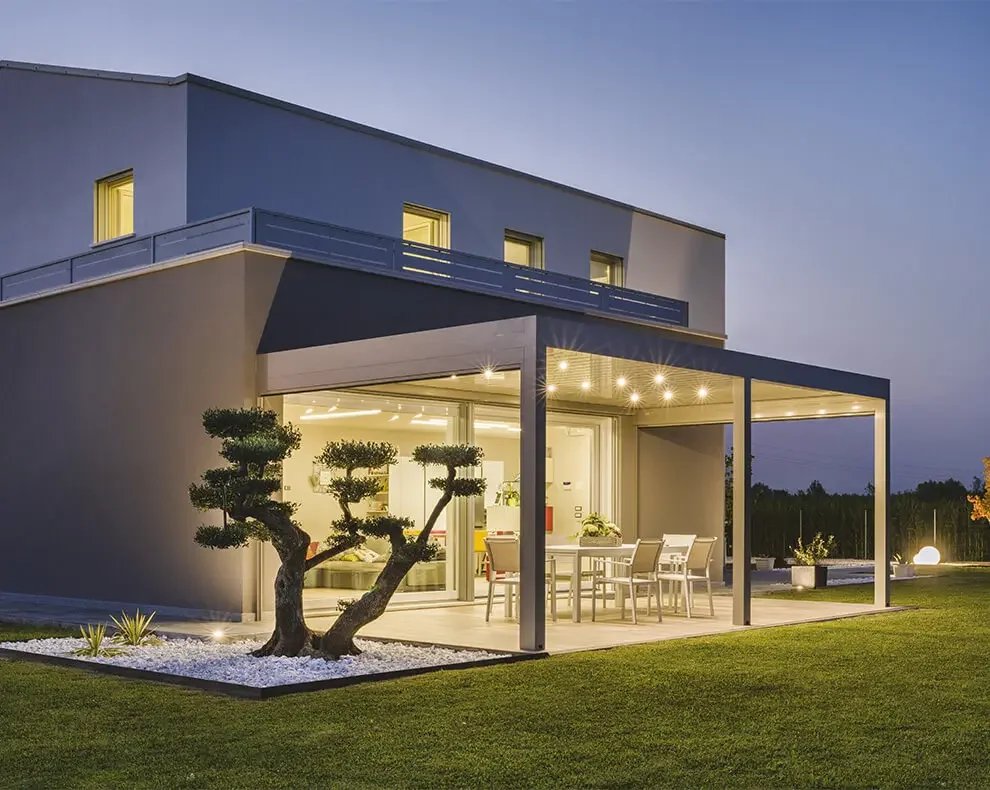 Modern house with a lit outdoor patio, glass sliding doors, and a neatly manicured lawn with a sculpted tree.