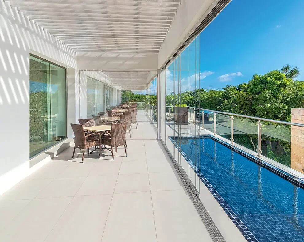 Balcony with outdoor dining tables and chairs, glass railing, small pool, and lush green trees in the background under a clear blue sky.