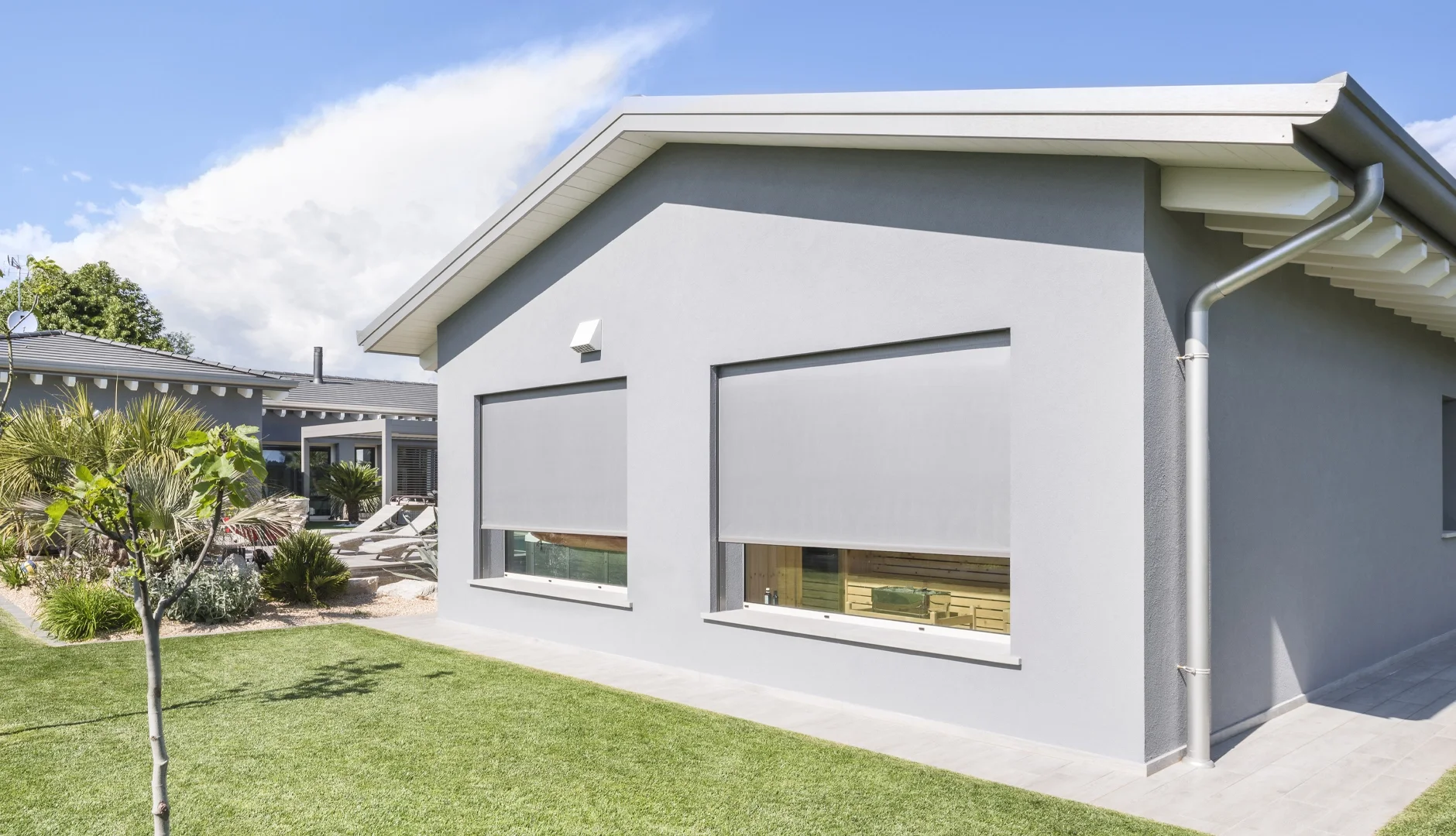 Front view of a modern gray house with two large windows and a sloped roof; well-maintained green lawn with small trees and shrubs; neighboring house visible in background; partly cloudy sky.