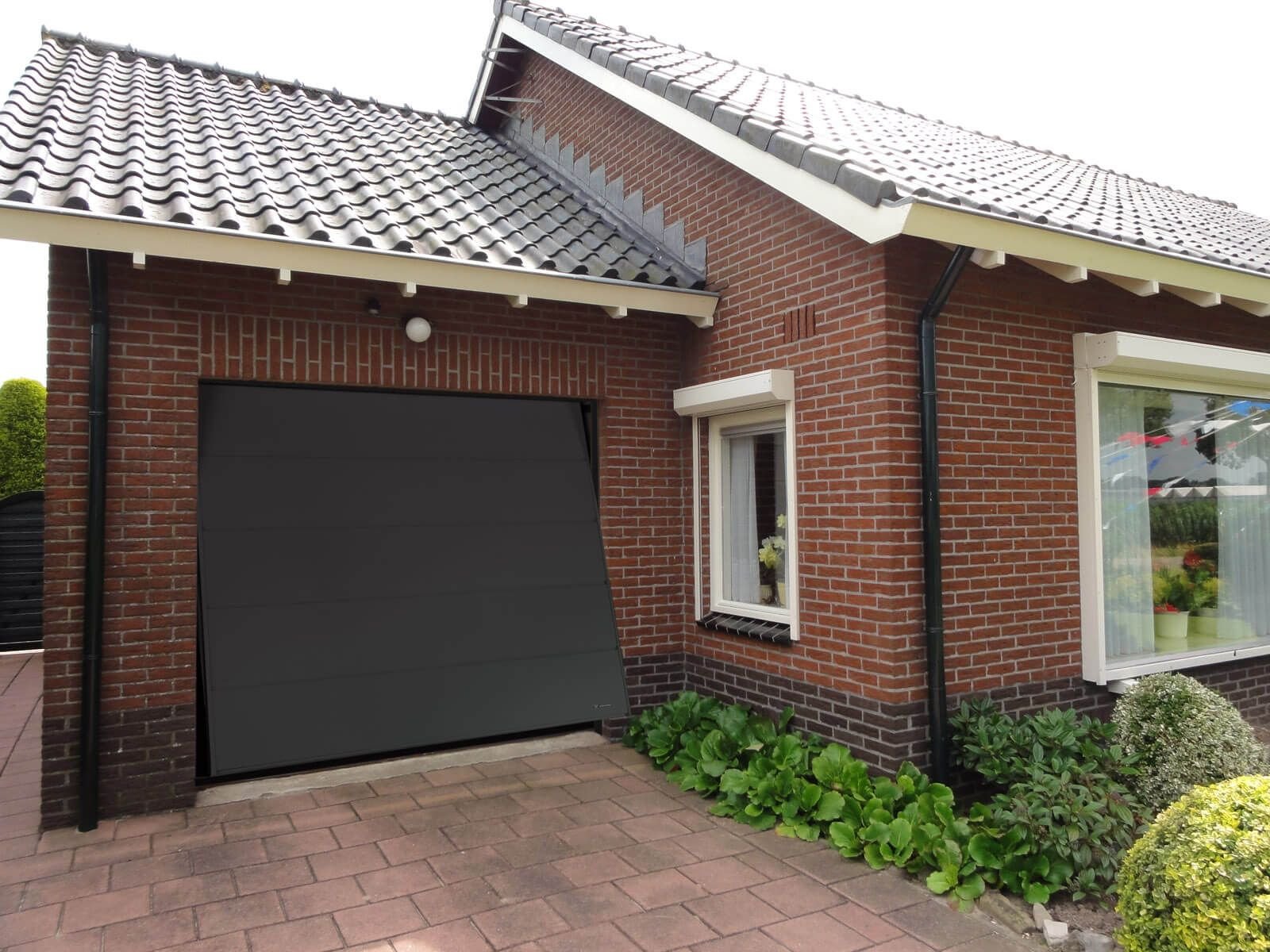 The image shows a brick house with a closed black garage door and a small window with curtains. There are green plants and bushes near the house, and the driveway is made of red brick pavement.