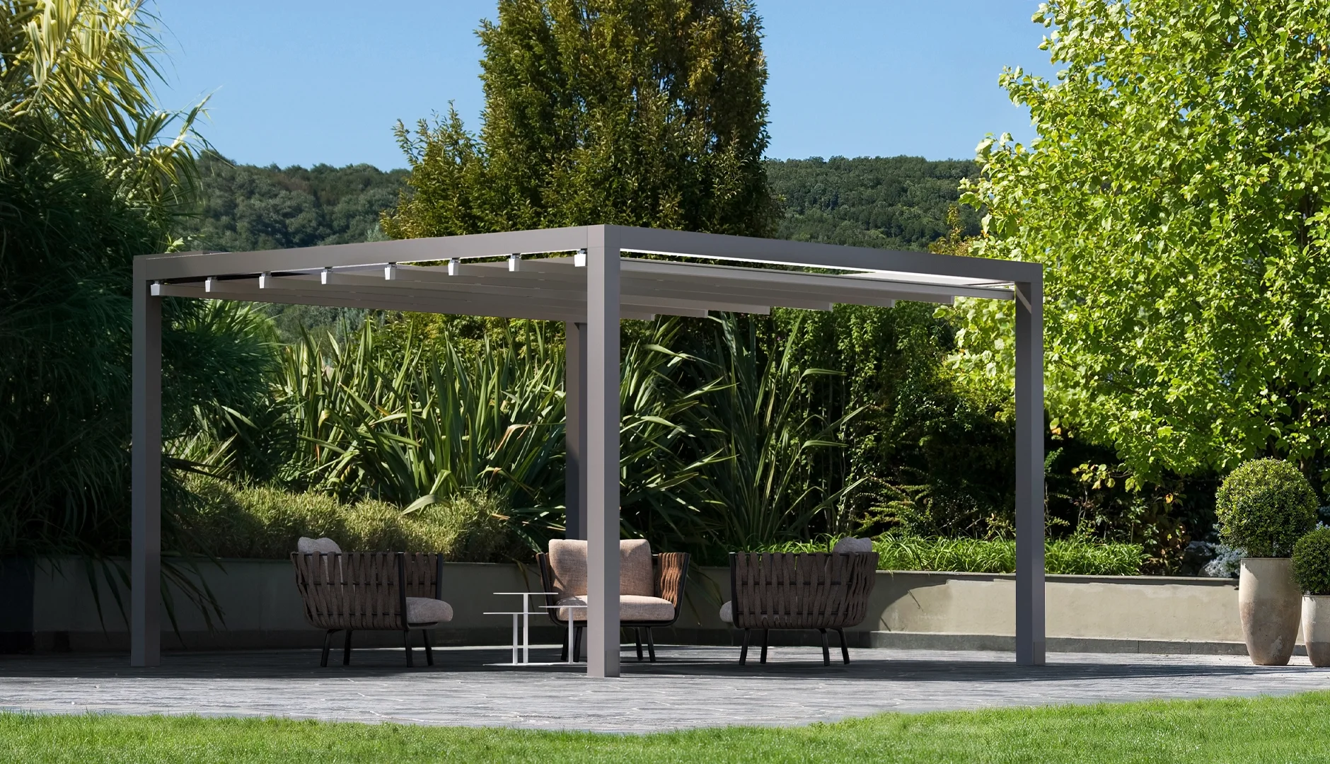 An outdoor patio with modern furniture under a pergola, surrounded by lush greenery and potted plants, with mountains in the background on a sunny day.