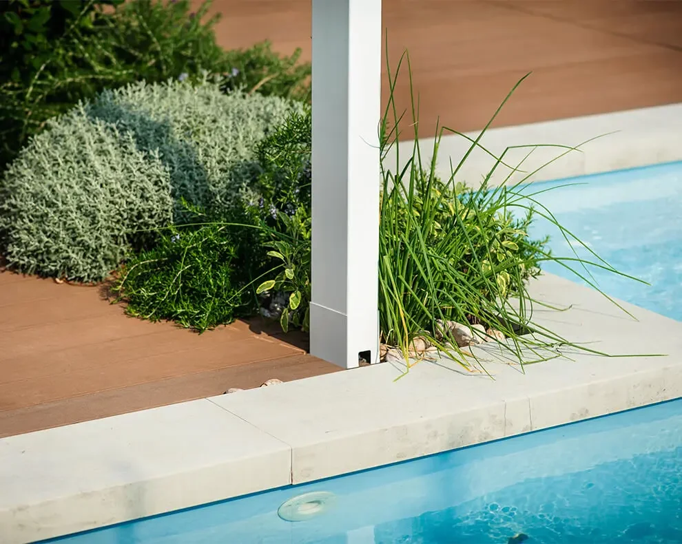 A close-up of the edge of a swimming pool with green grass and plants beside a wooden deck, partially separated by a white post.