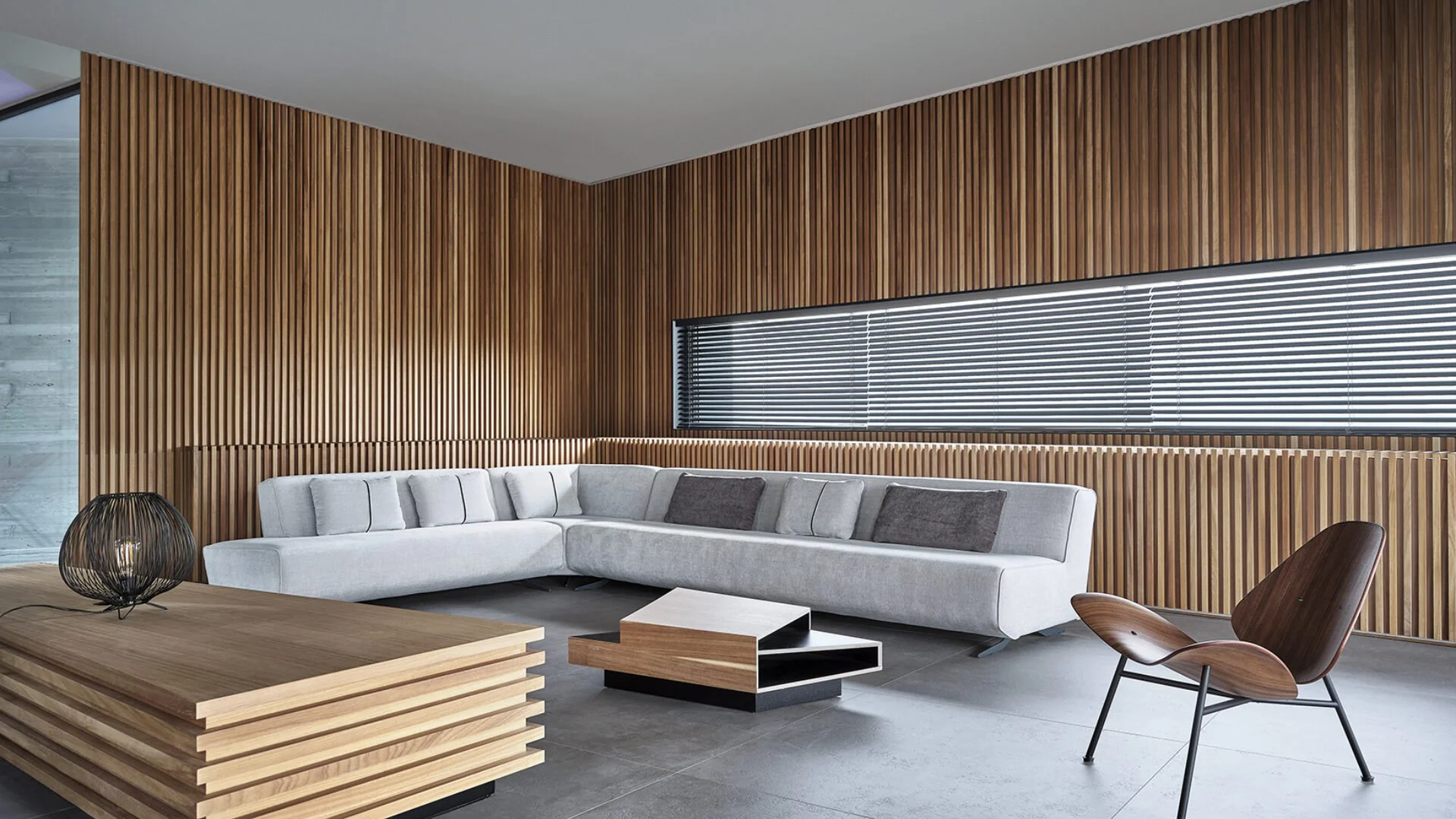 Modern living room with wooden slat wall, a white sectional sofa with pillows, a wooden coffee table, a stylish wooden armchair, and horizontal window blinds.
