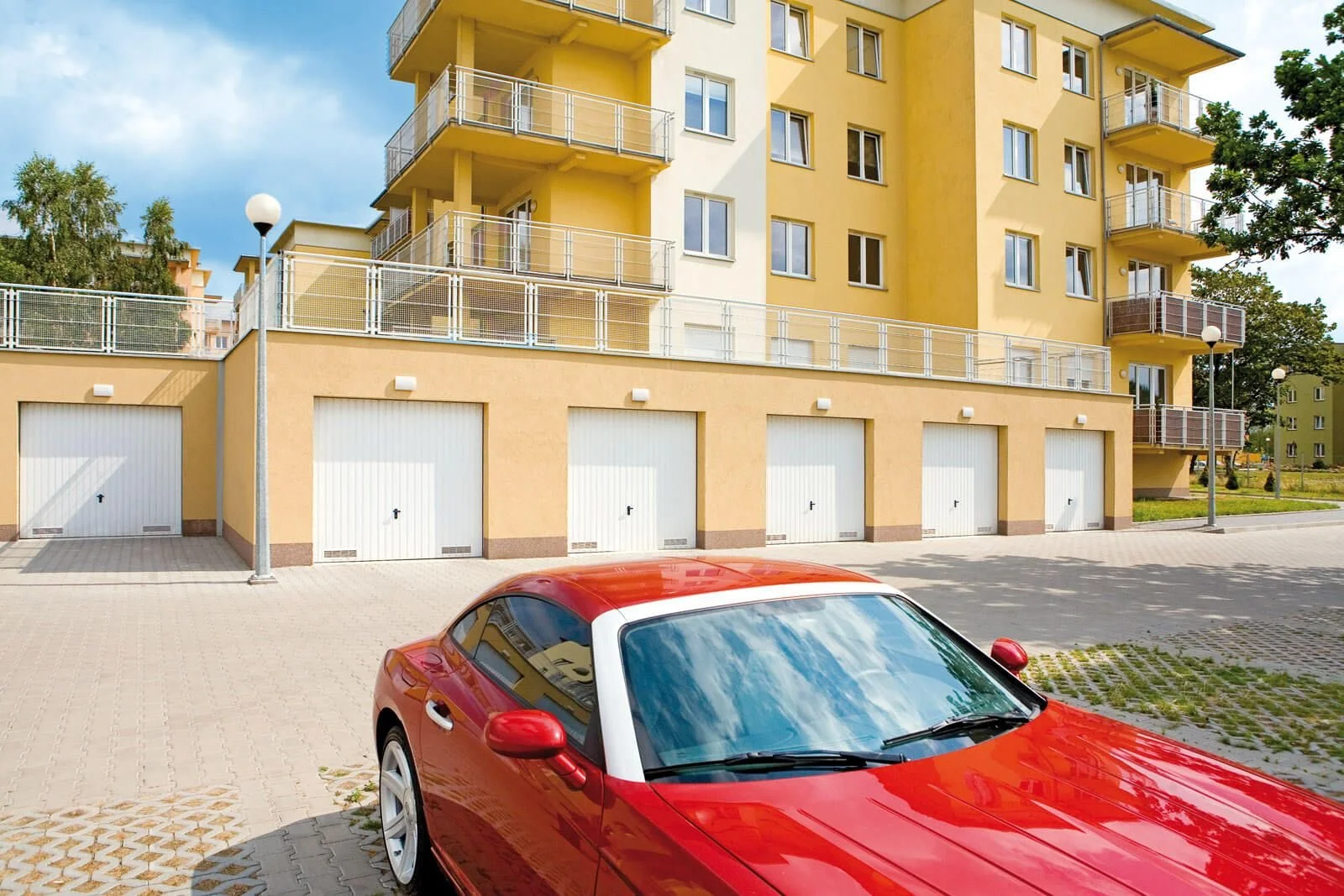 A red sports car parked in front of a modern yellow apartment building with white garage doors and balconies, under cloudy sky.