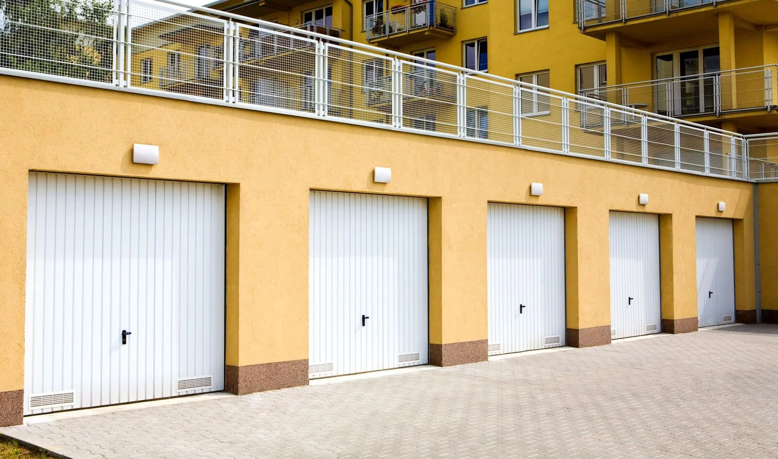View of a yellow residential building with four white garage doors at ground level and a paved area in front. The upper levels have balconies with metal railings.