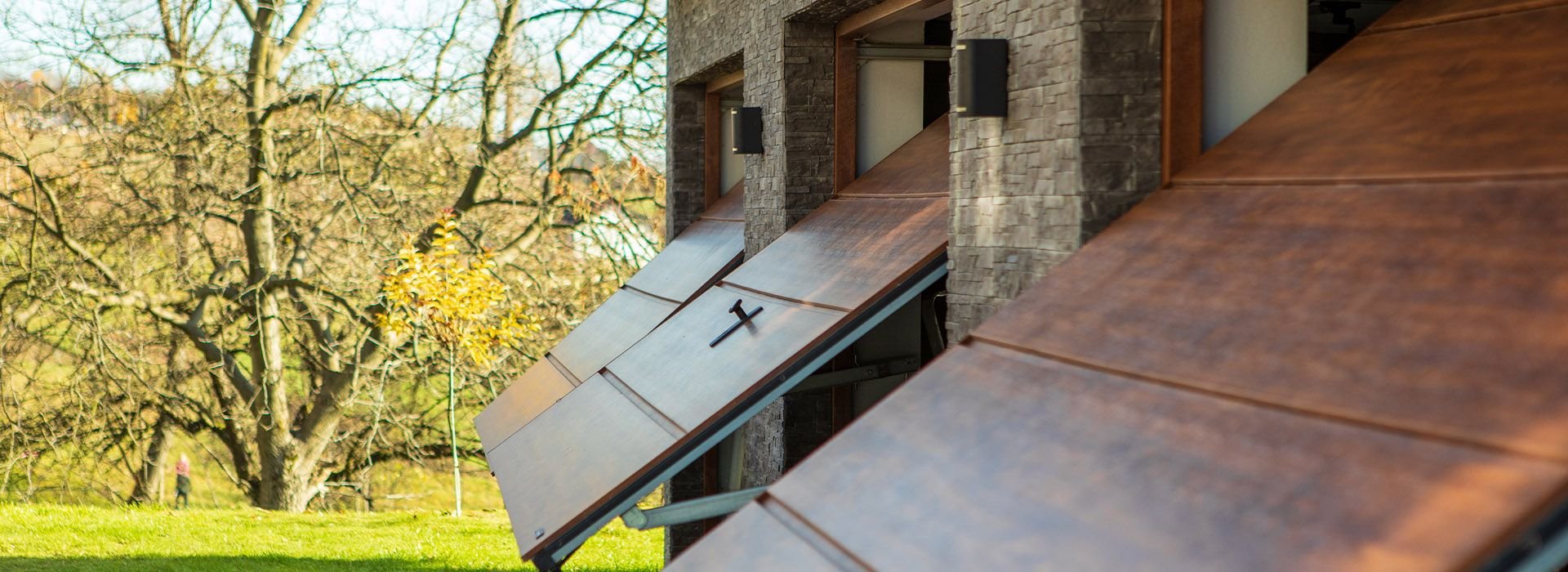Close-up of a row of solar panels installed on a sloped roof of a building, with a background of leafless trees and another tree with yellow leaves.