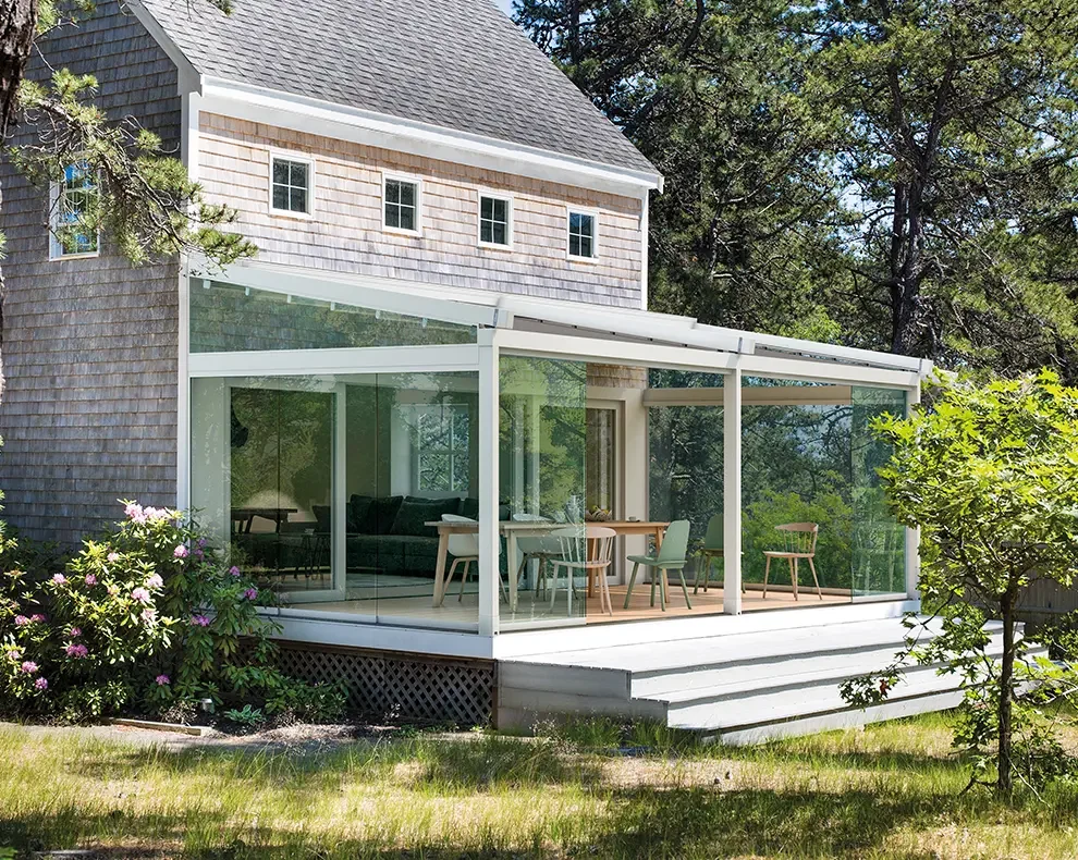 A house with an enclosed glass porch and outdoor stairs leads to a grassy yard with trees and flowering bushes.