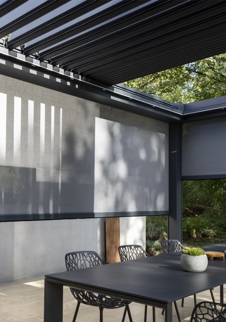 Modern outdoor patio with a black table, contemporary chairs, a white planter with greenery, a concrete wall, and a shade with sunlight filtering through trees.
