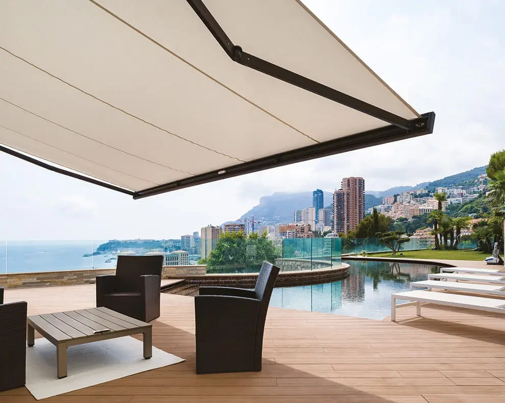 Outdoor terrace with a large beige retractable awning, black wicker chairs, a small wooden table, a pool, lounge chairs, and a cityscape with mountains in the background.