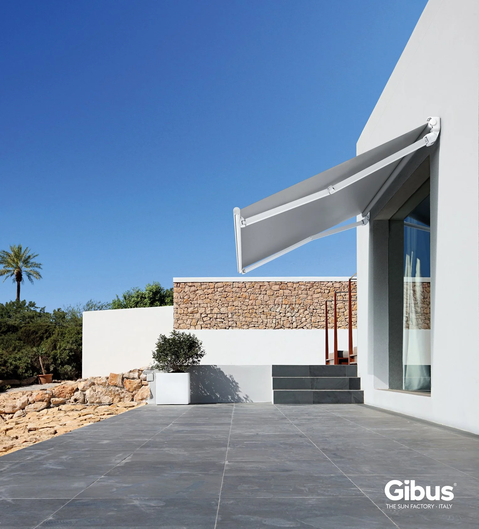 Modern house exterior with a large window, a white wall, a stone wall, tiled patio, potted plant, and a retractable awning, under a clear blue sky.