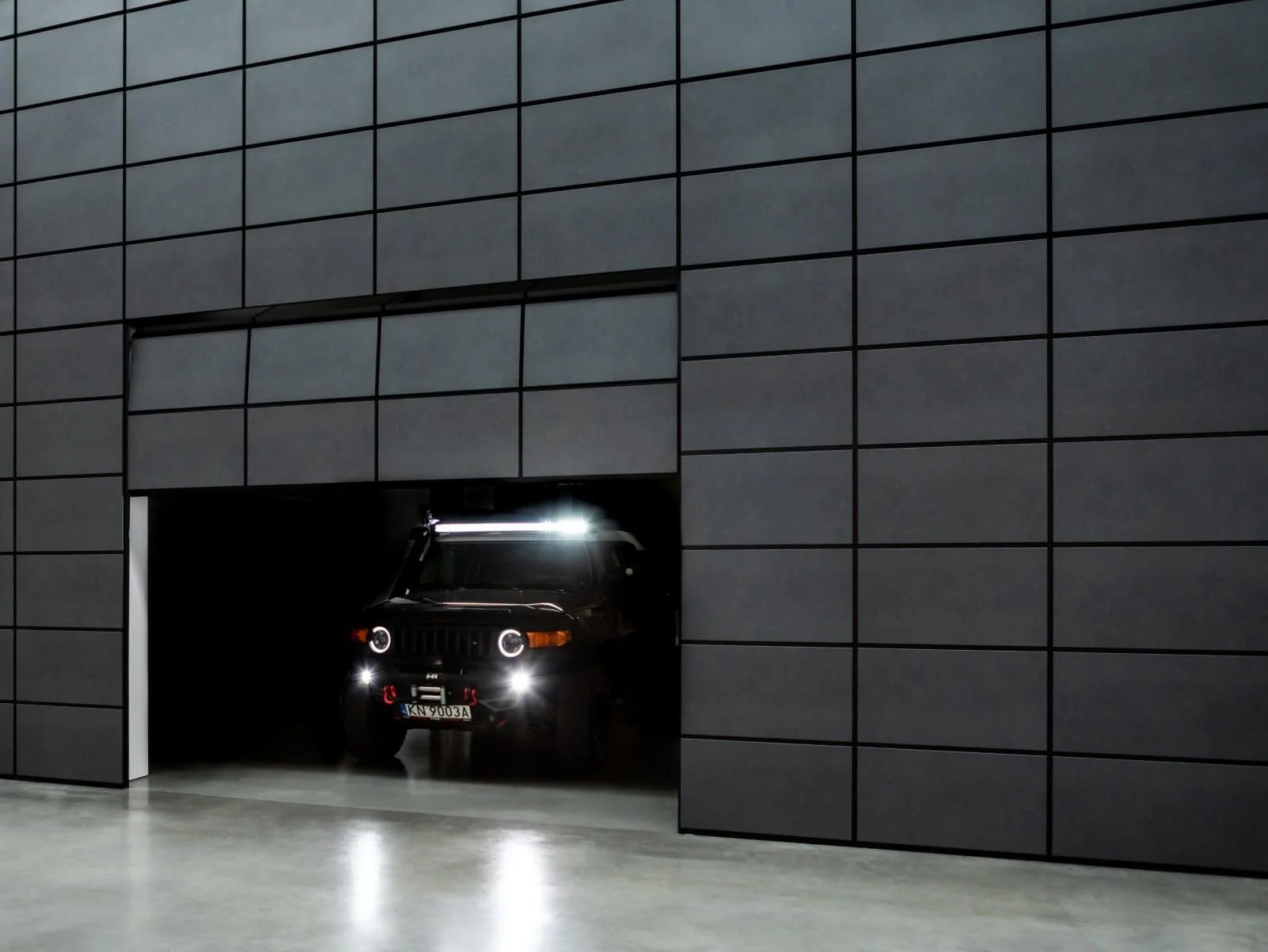 A black SUV partially visible as it exits a dark garage through an opening in a sleek, modern building with a gray tiled exterior.