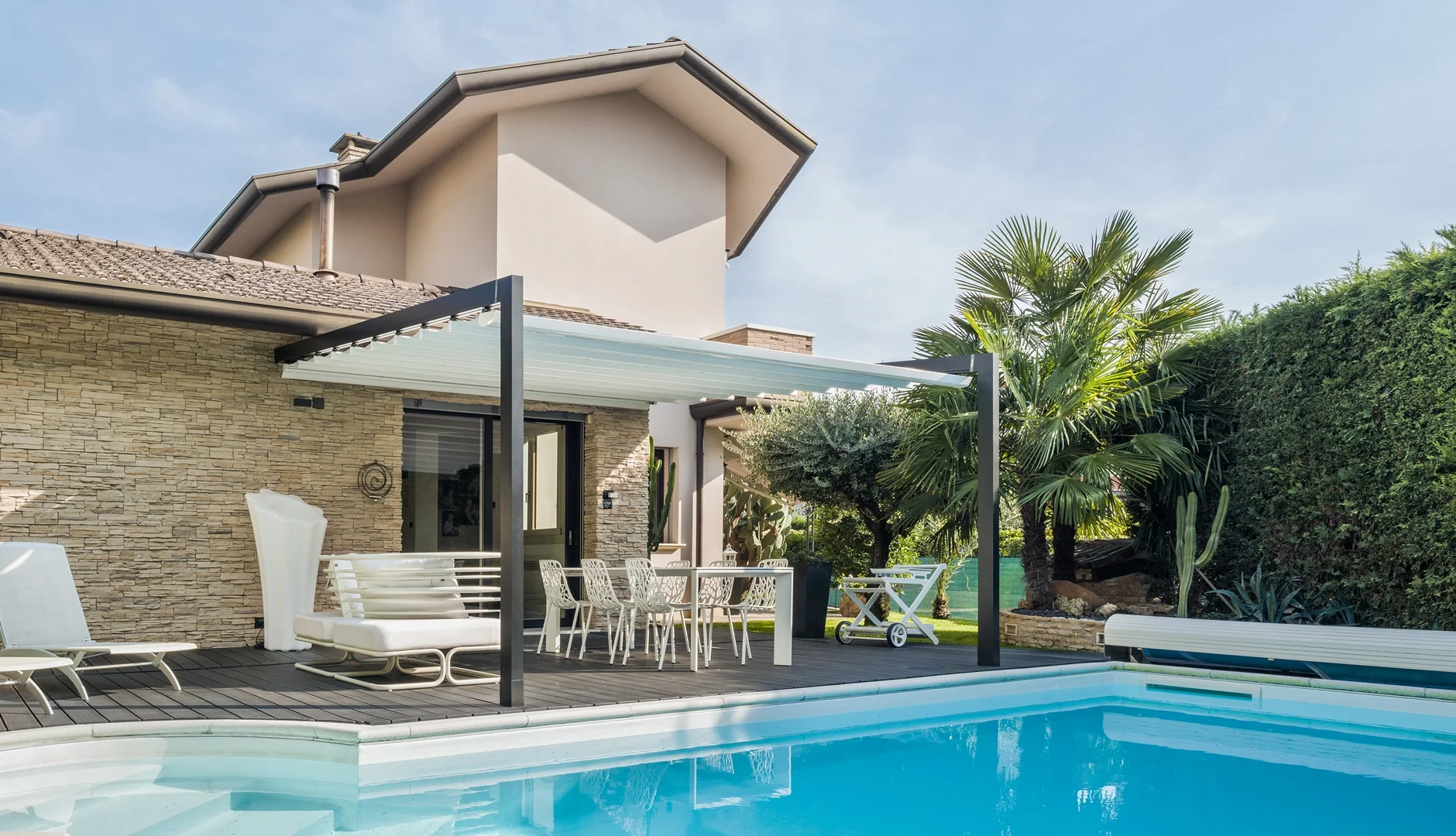 Luxury backyard with a swimming pool, lounge chairs, outdoor dining area, lush green plants, palm trees, cacti, and a modern house with a stone wall and a retractable awning.