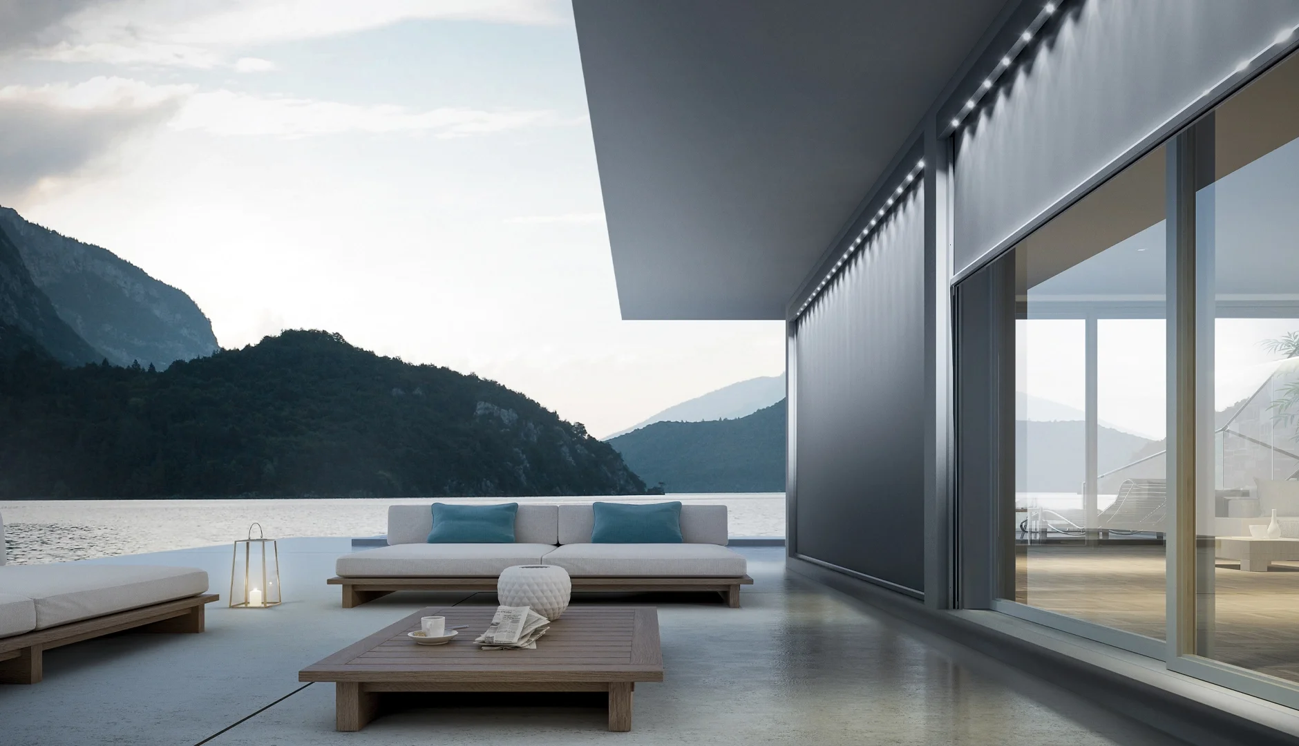 Modern outdoor patio with a view of mountains and water, featuring white sofas with blue pillows, a wooden coffee table with a newspaper and a coffee cup, a lantern, and glass doors leading inside a contemporary house.