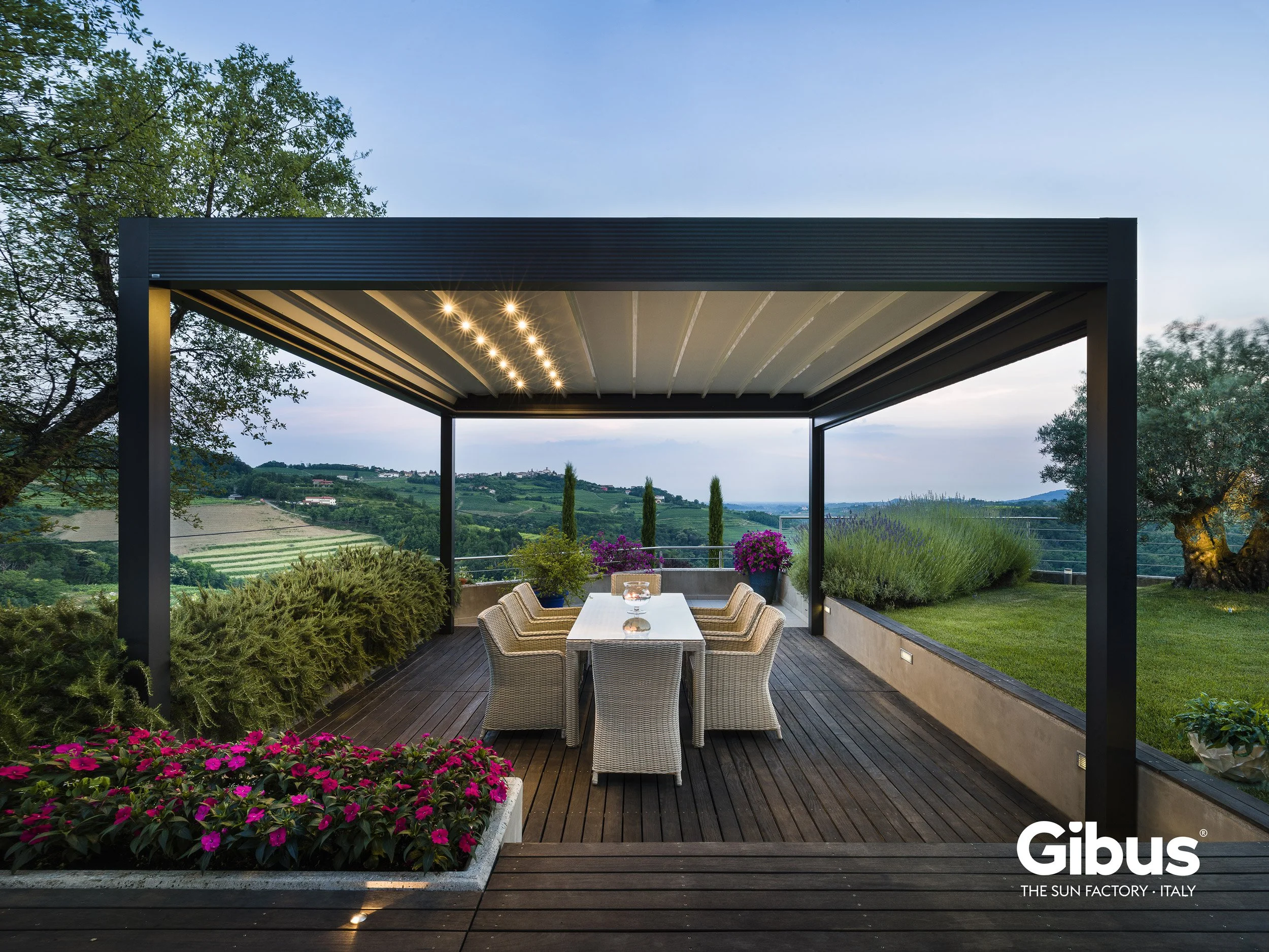 Outdoor terrace with a table and chairs, surrounded by plants and overlooking a scenic landscape of hills and vineyards during dusk.
