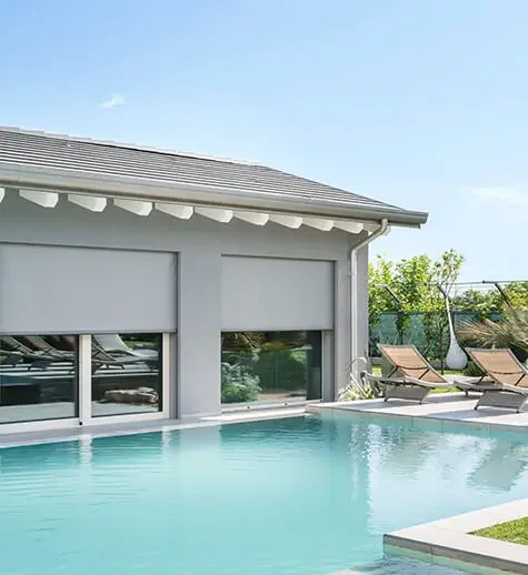 Swimming pool with two lounge chairs outdoors beside a modern house with large windows and a gray roof under a partly cloudy sky.