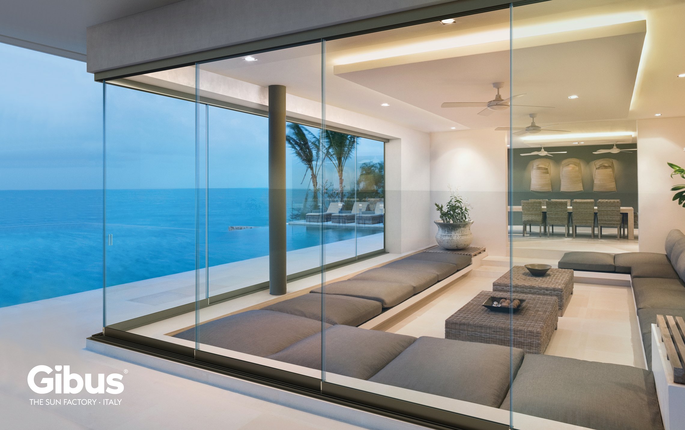Modern living room with large glass windows overlooking the ocean, outdoor pool, and palm trees, featuring gray cushions, wicker tables, potted plants, and a dining area in the background.