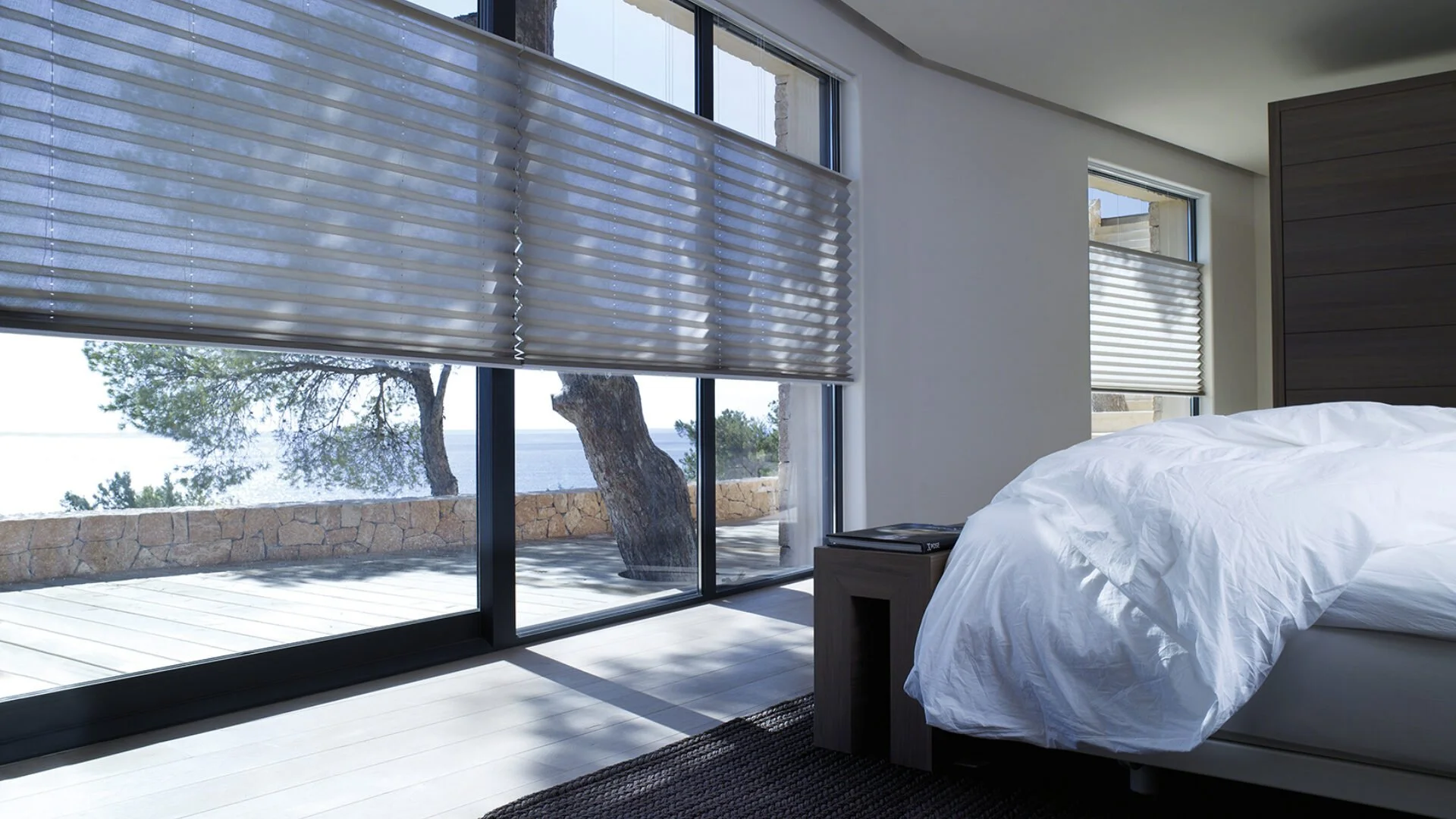 Interior of a bedroom with large glass sliding doors leading to a balcony, view of trees and water outside, white bed with crumpled bedding, small wooden nightstand, and window blinds.