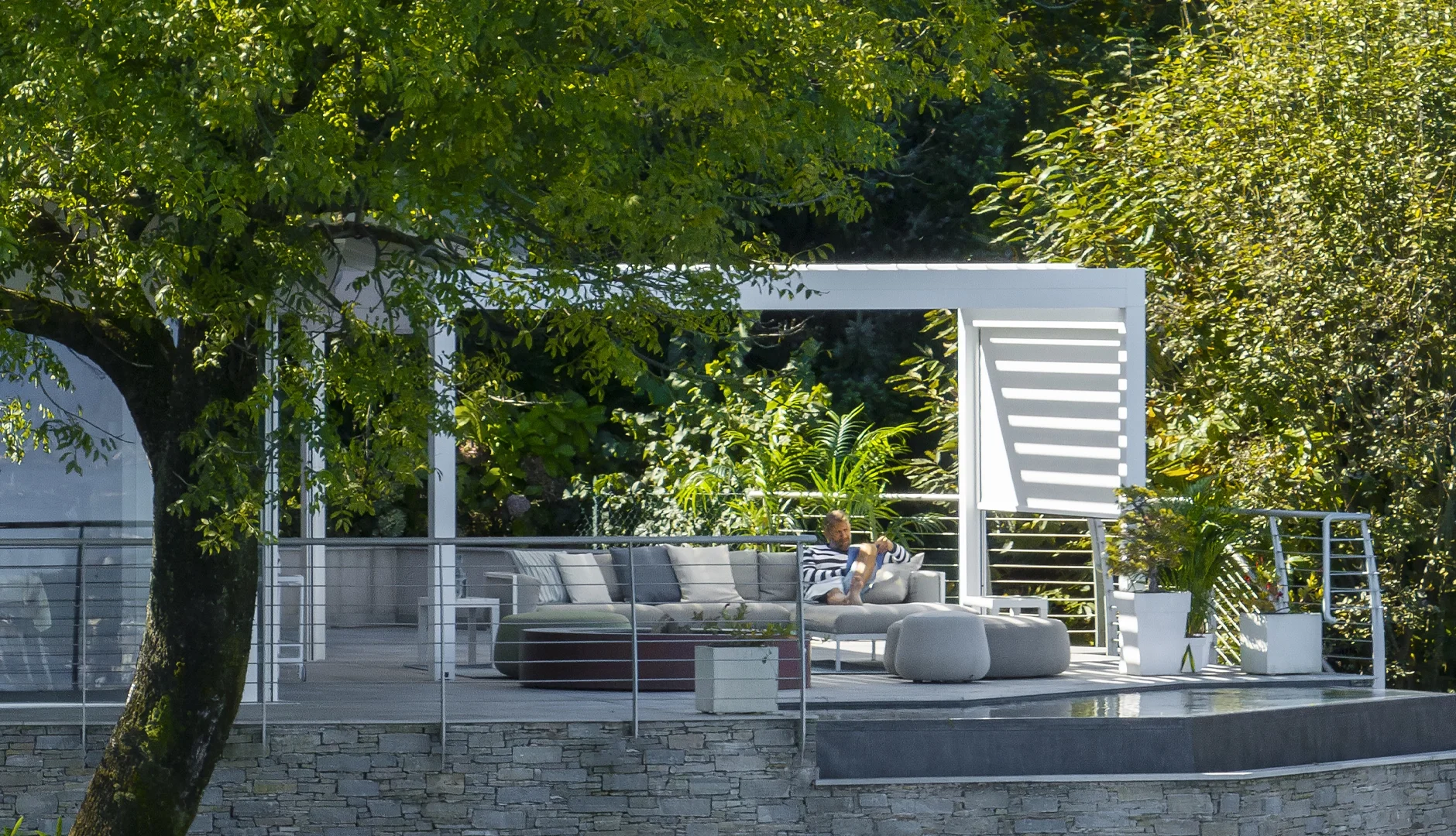 A person relaxing on a white outdoor lounge on a modern patio with greenery and trees around.