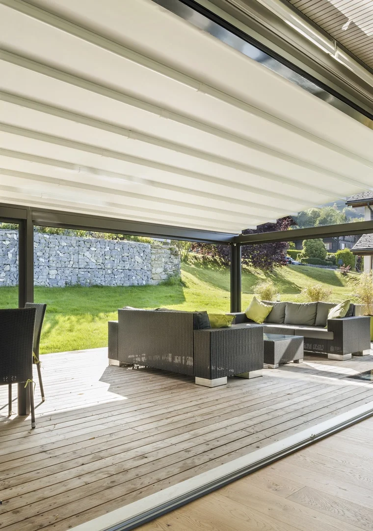 Outdoor patio with wicker furniture including a sectional sofa with cushions, on a wooden deck, view of a grassy yard, stone wall, and trees beyond.