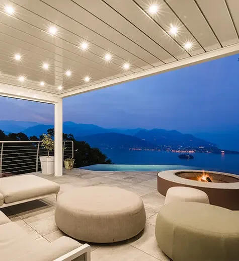 Modern outdoor patio with cushioned seating, a fire pit, and a scenic mountain and water view at dusk.